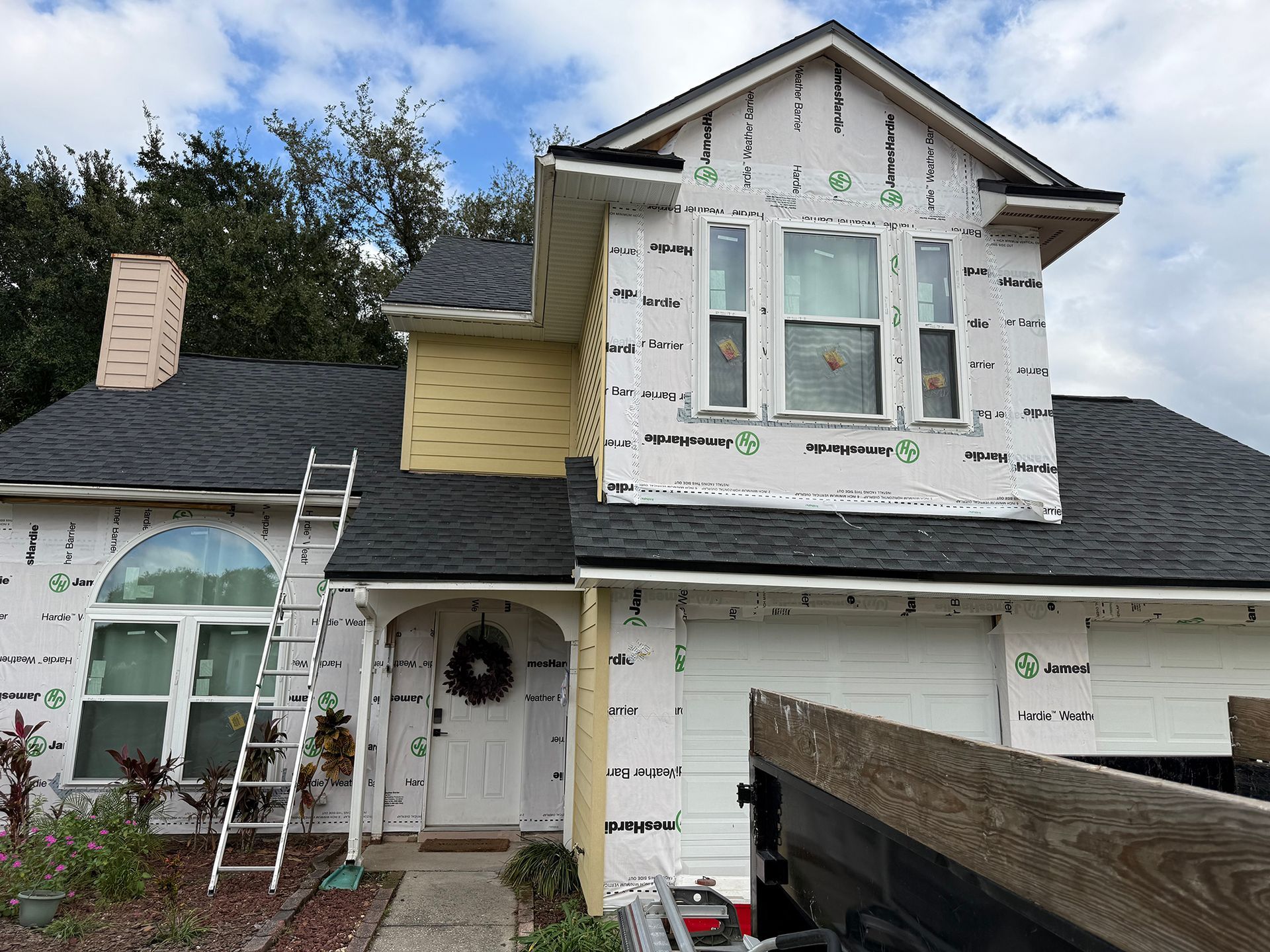 Partially renovated two-story house with white siding, ladder, and scaffolding under a cloudy sky