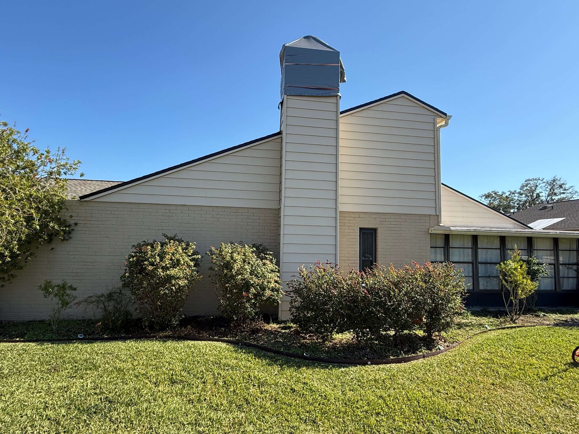 Suburban house exterior with beige siding, central chimney, shrubs, and a green lawn under a blue sky