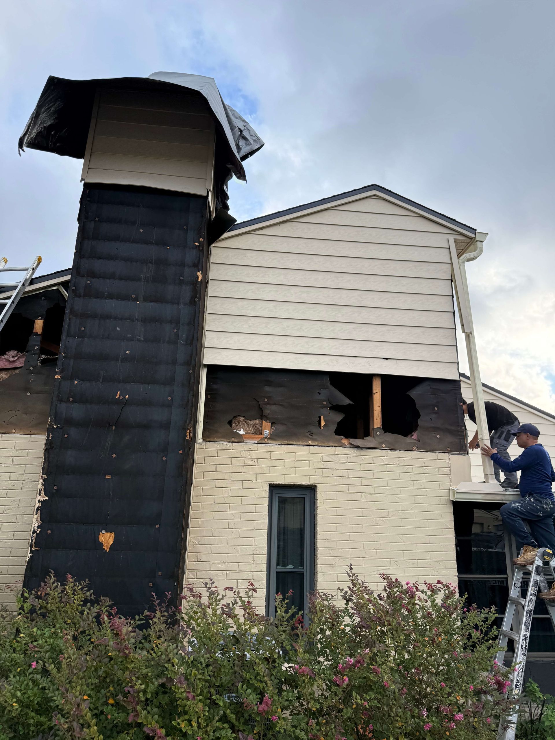 Fire-damaged house with blackened chimney and broken windows, person on ladder inspecting the exterior