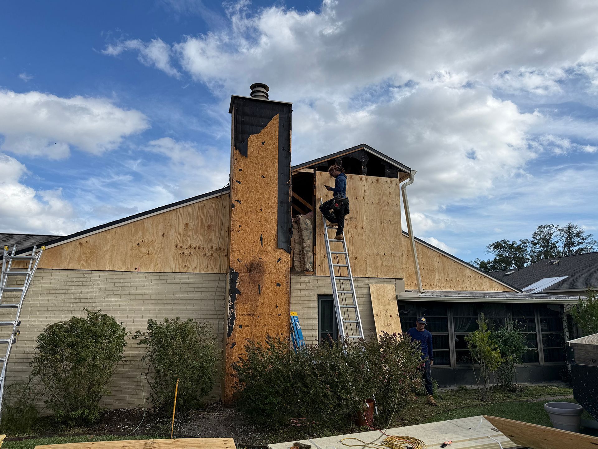 Damaged house roof with exposed plywood and a ladder under a cloudy sky