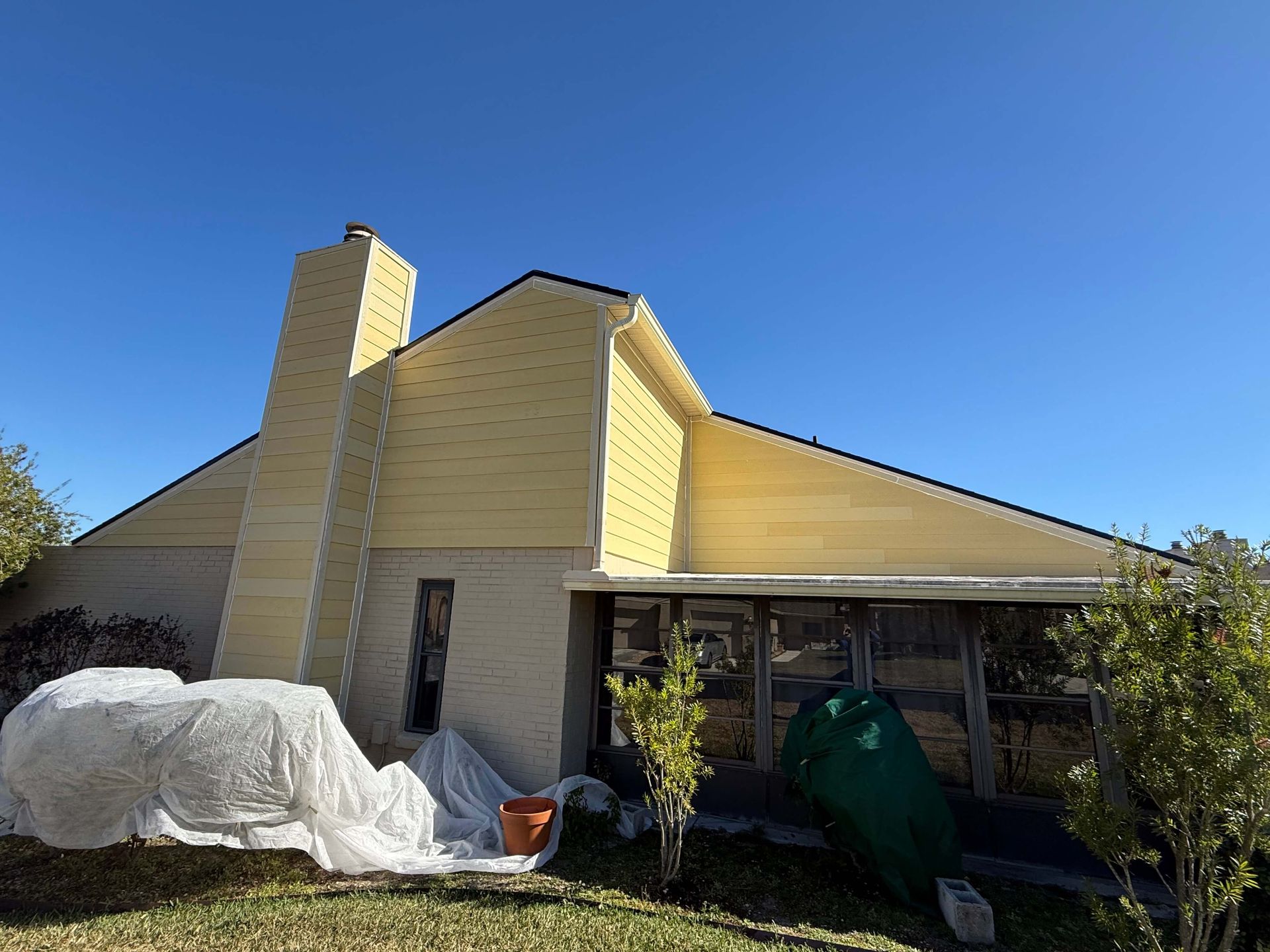 Small yellow house with a chimney, covered items in front, and a carport under a clear blue sky