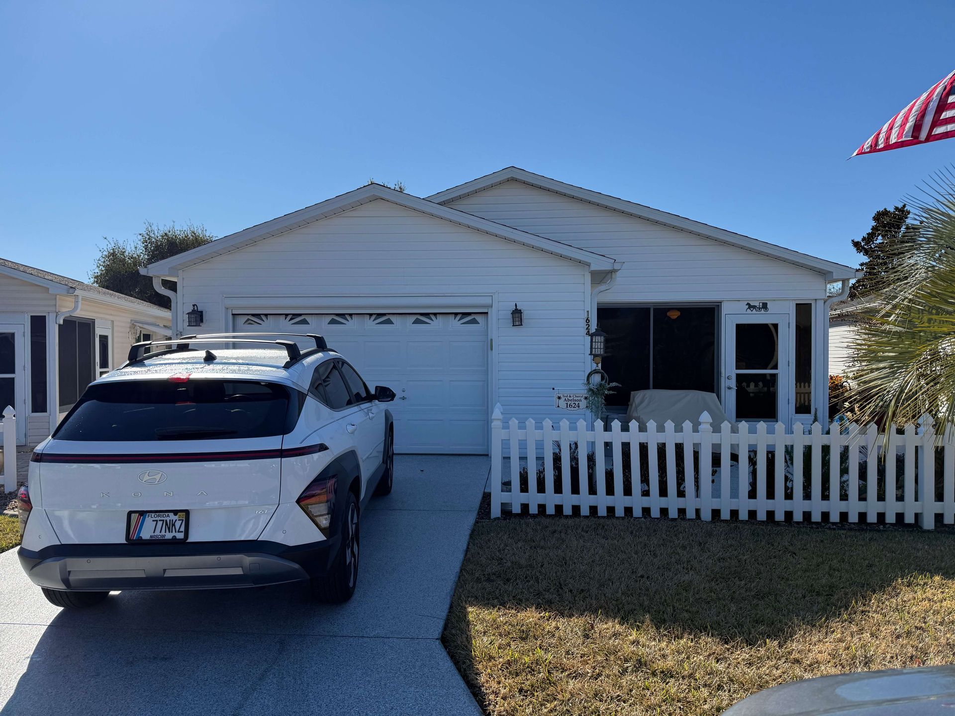 White SUV parked in a driveway outside a single-story house with a white picket fence and open garage.
