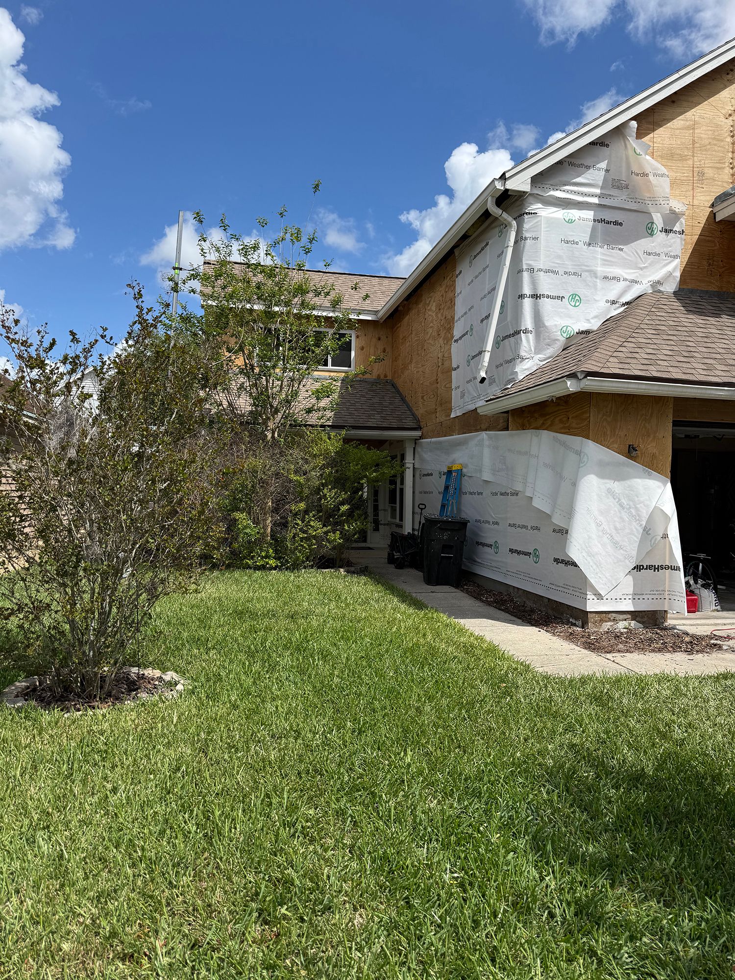 Suburban house exterior with green lawn, tree, and partly covered siding under a blue sky