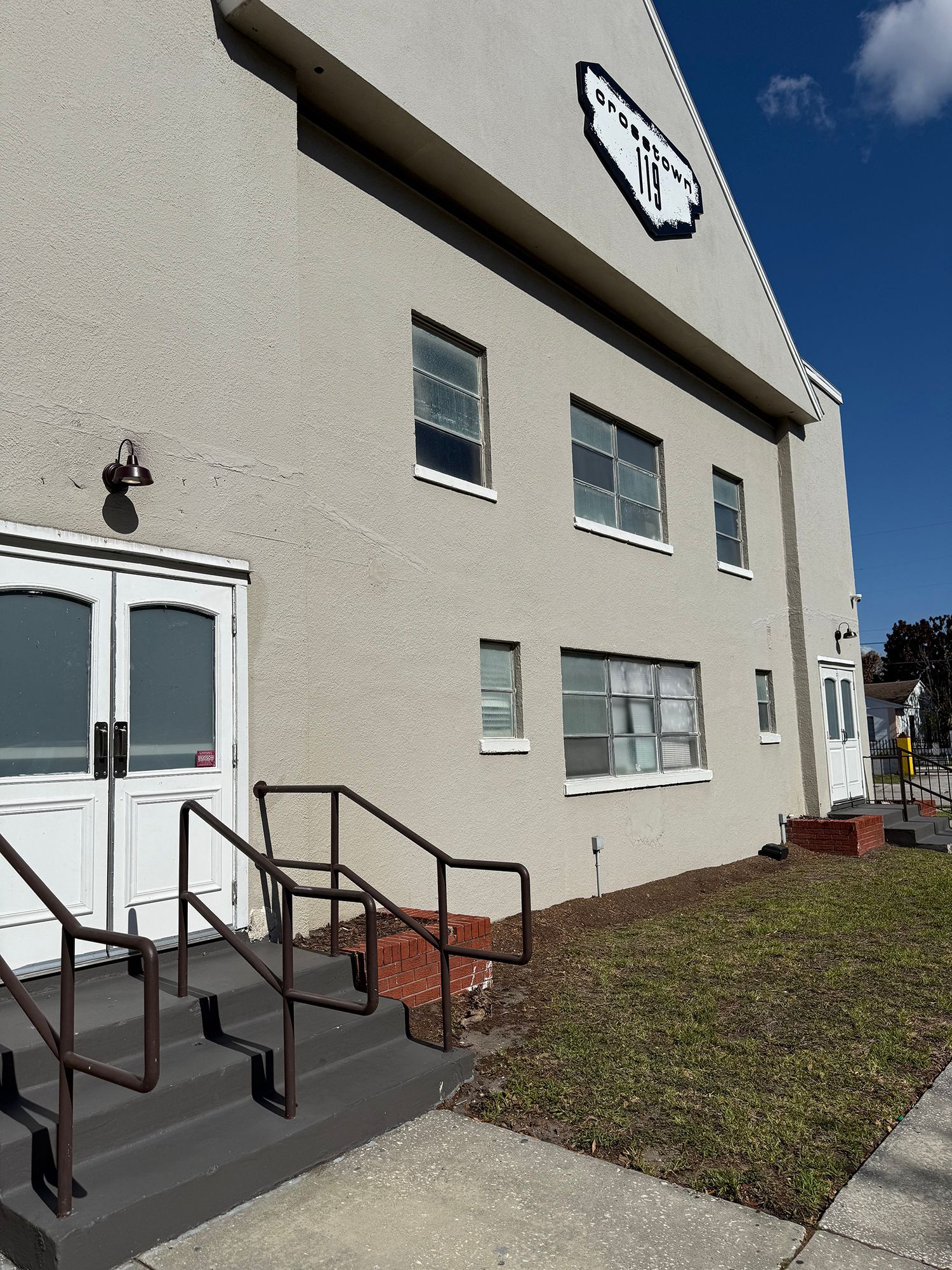Exterior of a beige building with a white sign, double doors, windows, and front steps with black railings.
