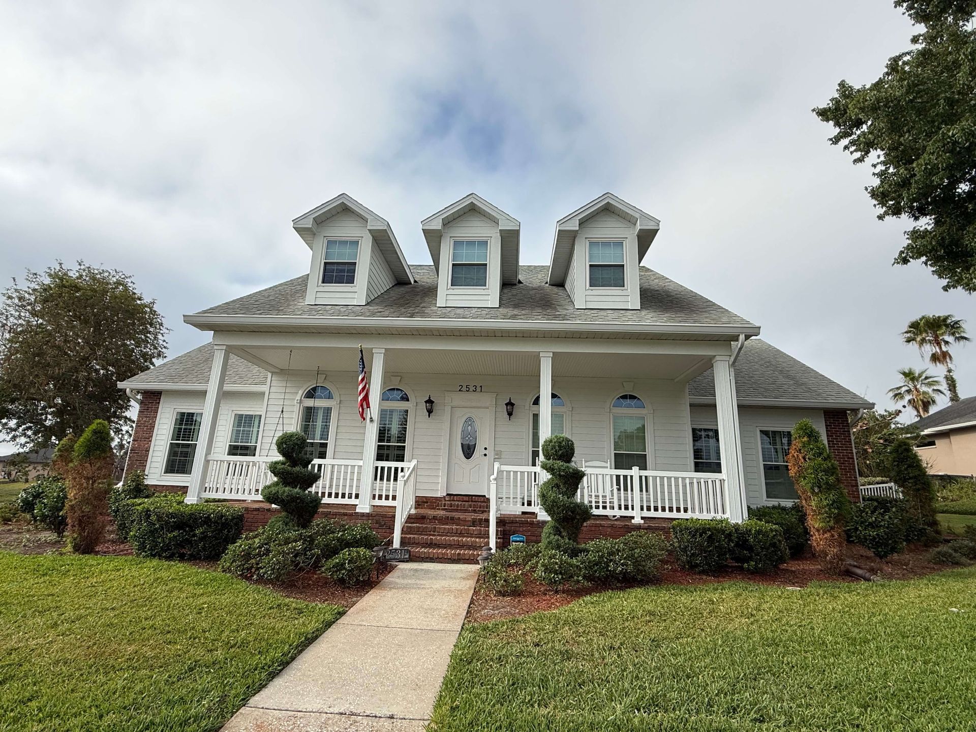 White house with a front porch, dormer windows, and brick steps, viewed from the lawn.