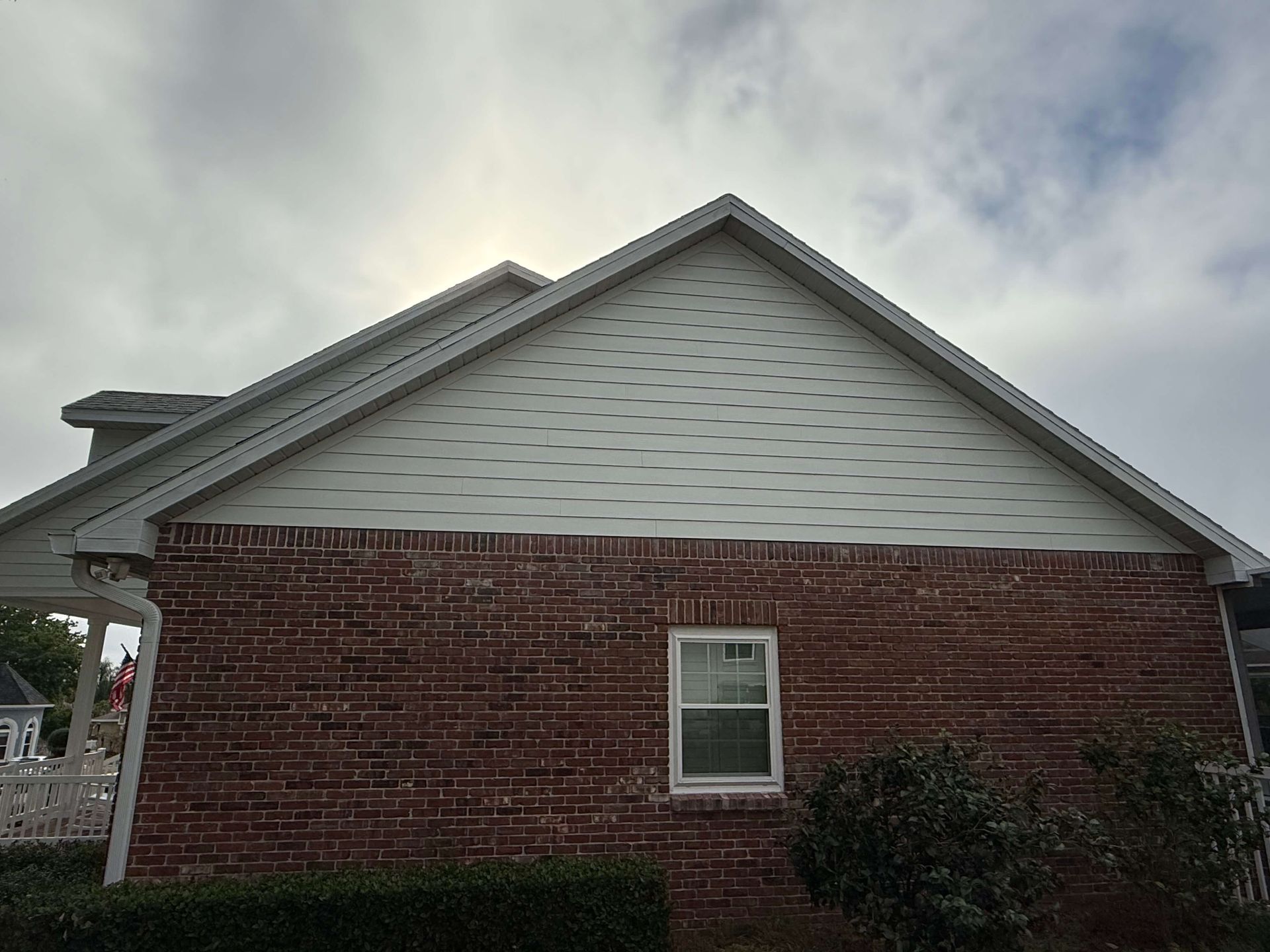 Red brick house side wall with white gable siding and a small window under an overcast sky