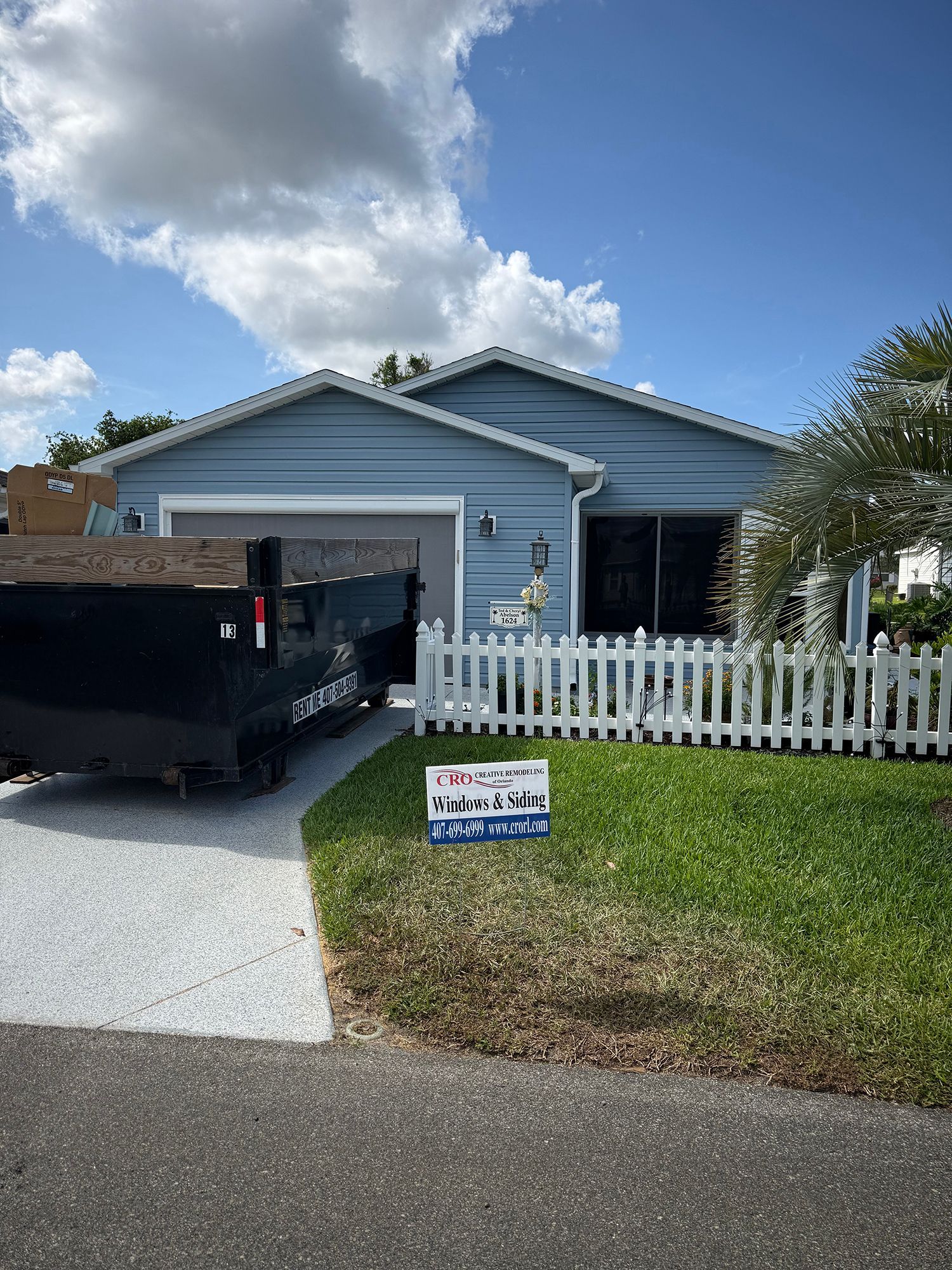 Blue house with open garage, black dumpster in driveway, and white picket fence under a cloudy sky.