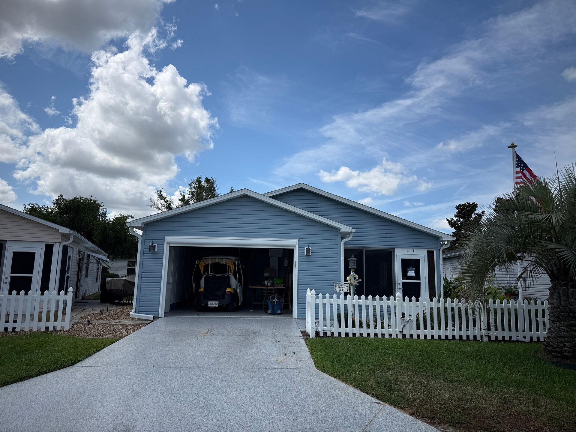 Blue single-story house with an open garage, white picket fence, and American flag under a cloudy sky