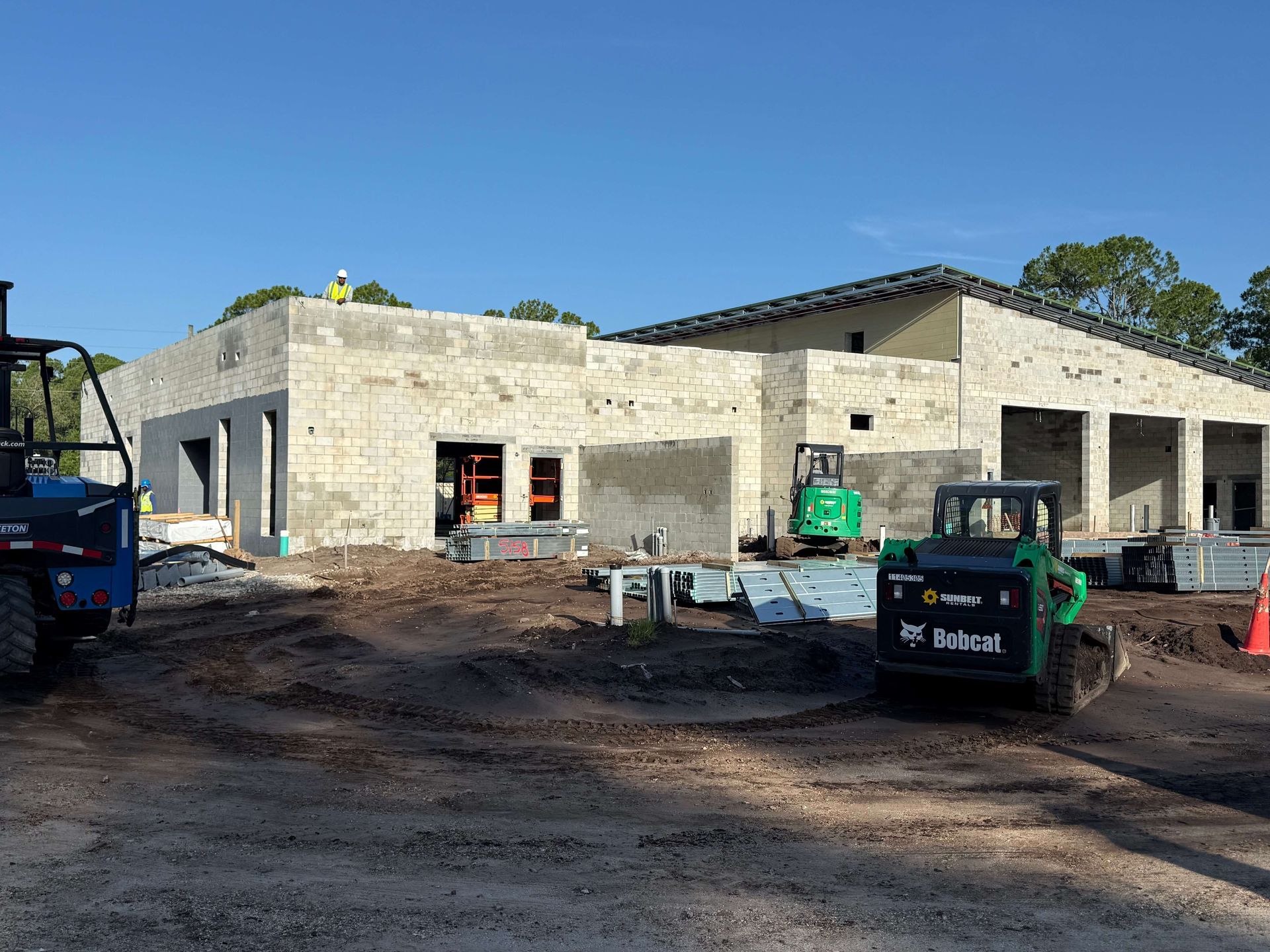Stone building under construction with exposed walls, roof framing, and equipment on a dirt lot.