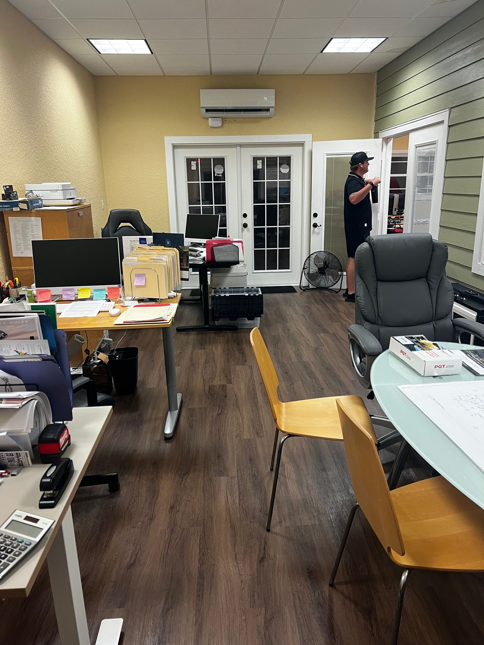 Office interior with desks, chairs, and a person standing near glass doors at the back.
