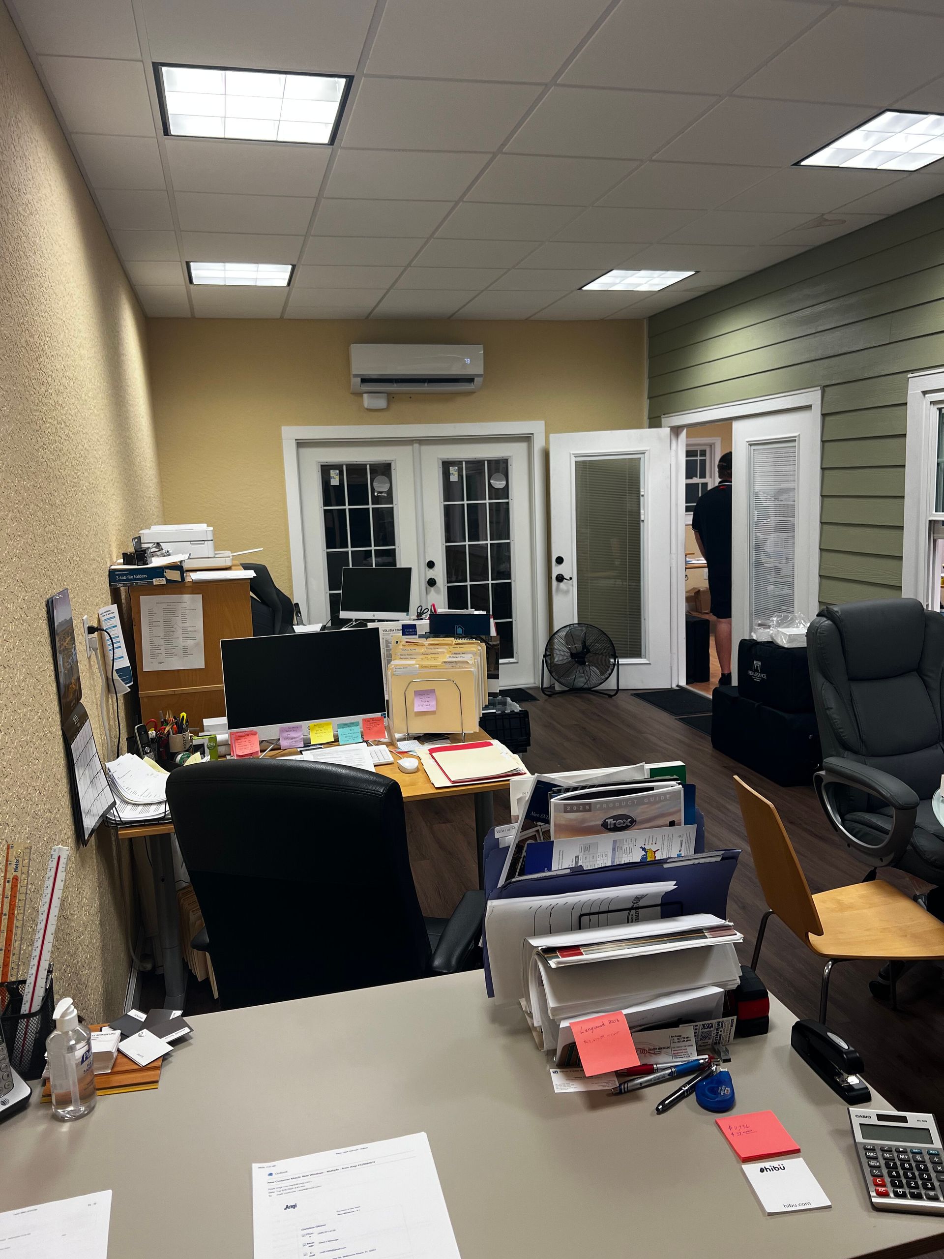 Cluttered office with desks, chairs, boxes, and a glass door at the back.