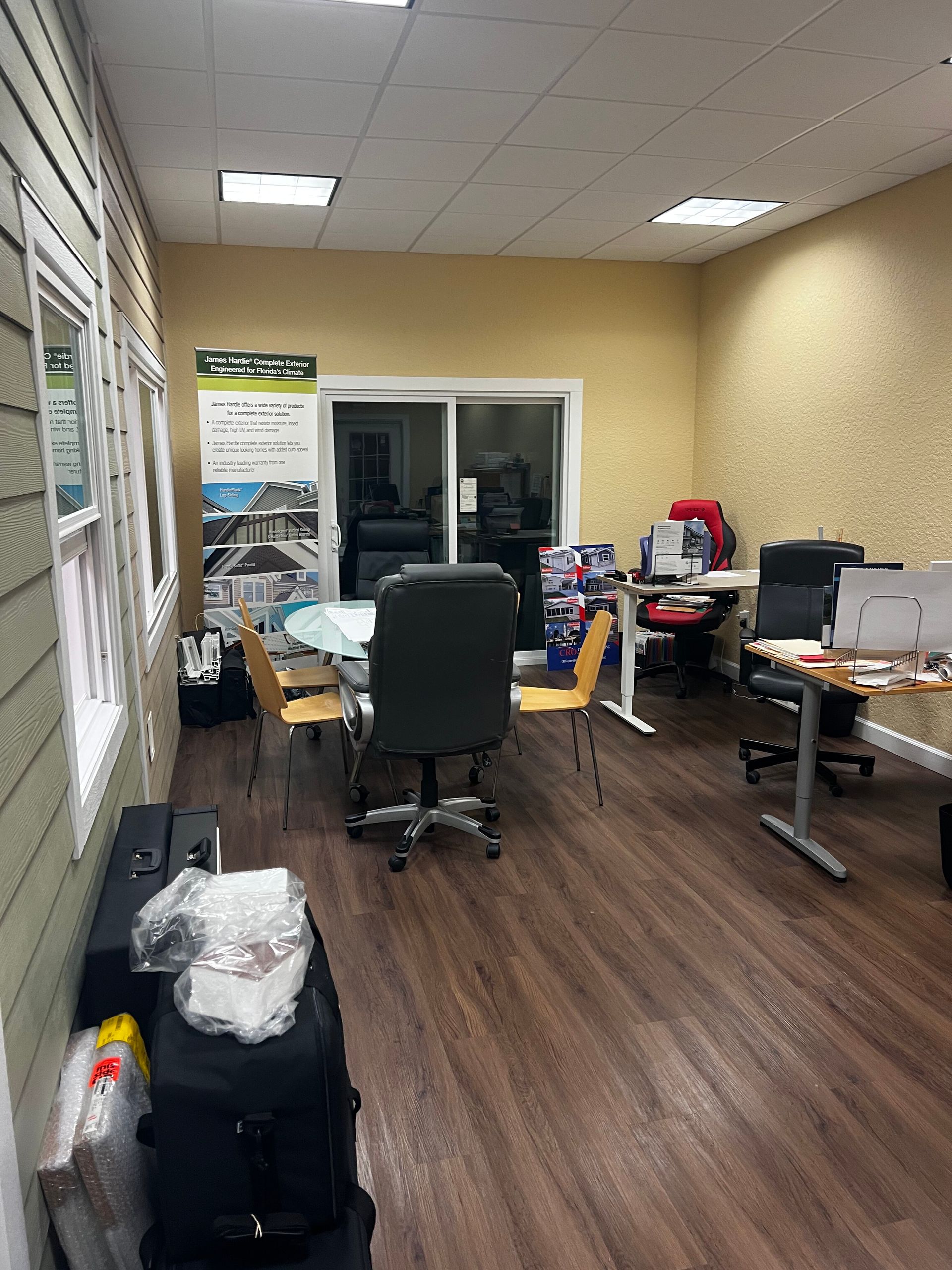 Small office with a desk, chair, printer, boxes, and wood flooring under fluorescent lights.