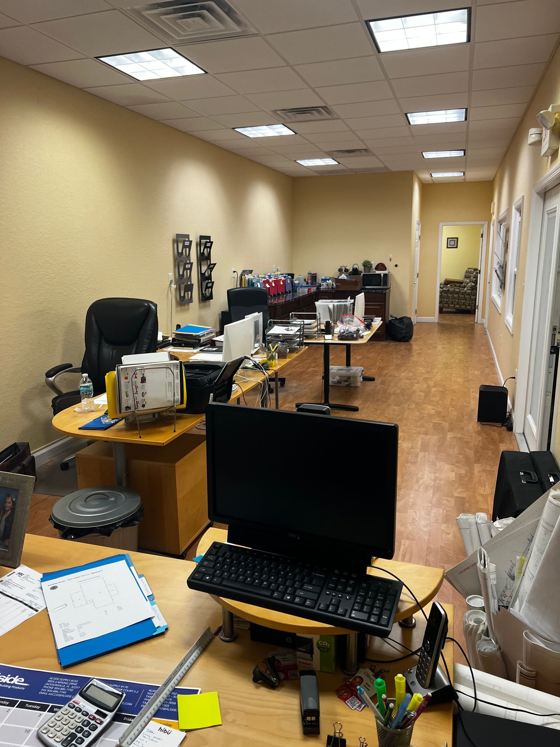Cluttered office with desks, computer monitors, papers, and boxes along a long hallway-like room.