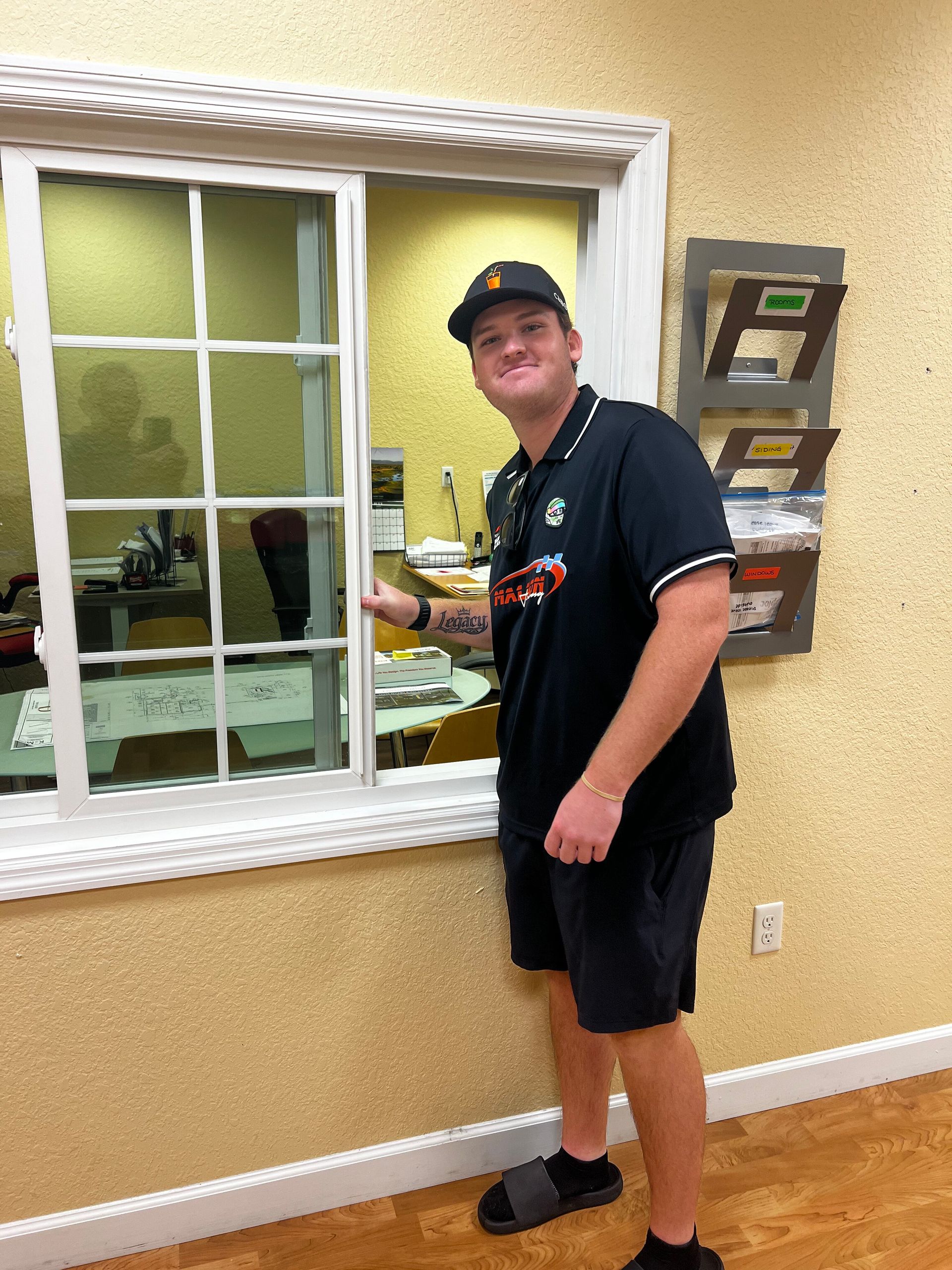 Man standing by a window in a yellow room, wearing a black cap, shirt, shorts, and slippers.