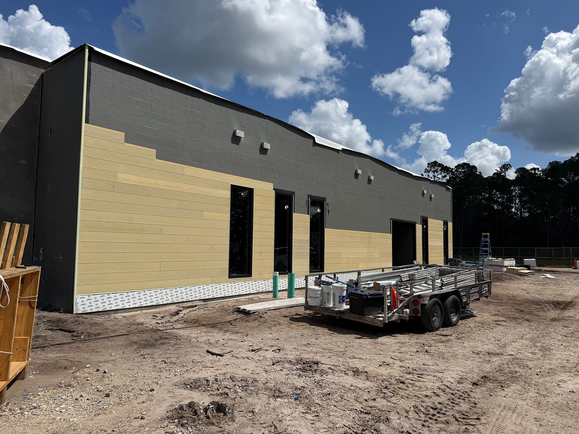 Partly finished tan commercial building with open doorways and construction trailer on a dirt lot under a cloudy sky