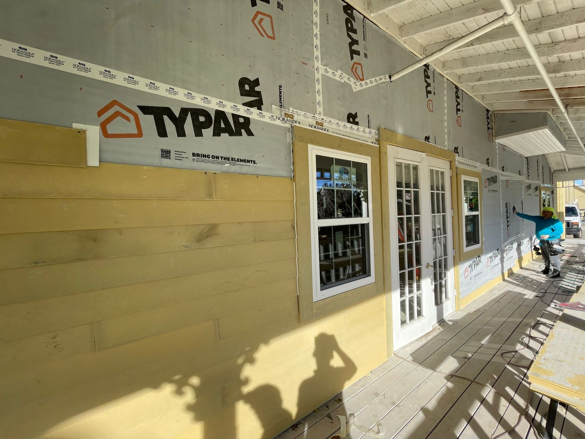 Partially finished building exterior with TYPAR wrap, windows, and a workbench on a covered porch