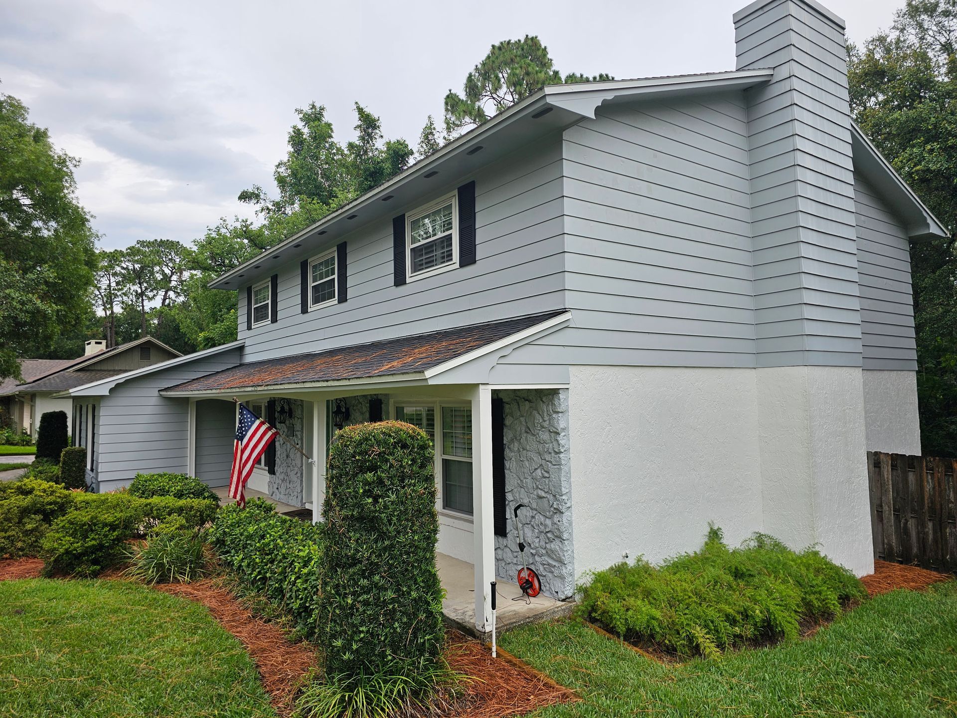 Two-story gray house with white trim, front porch, and an American flag, surrounded by greenery