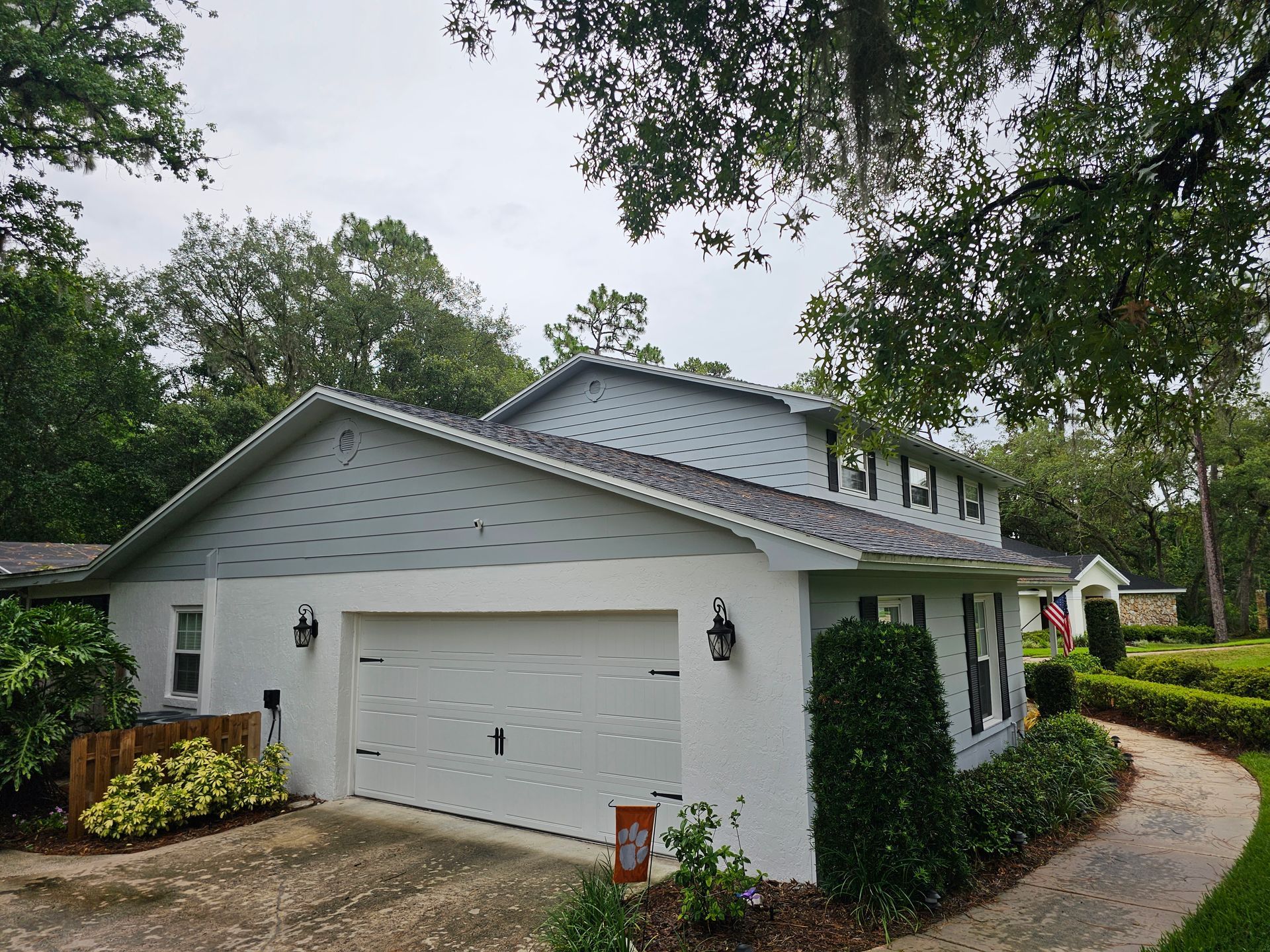 White two-car garage attached to a house with gray roof, surrounded by trees and shrubs.