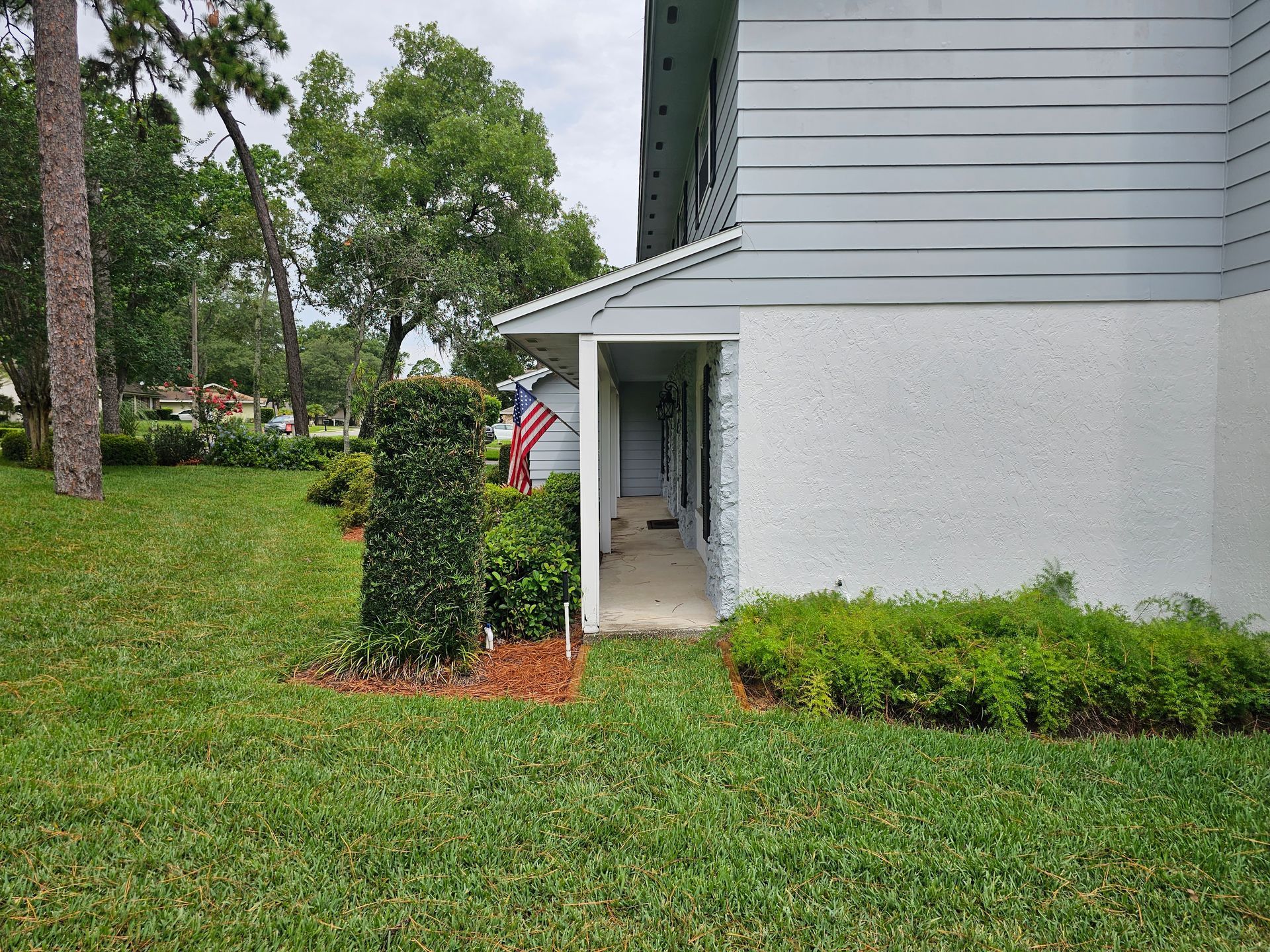 Side view of a gray house with a covered walkway, trimmed hedges, and an American flag by the entrance
