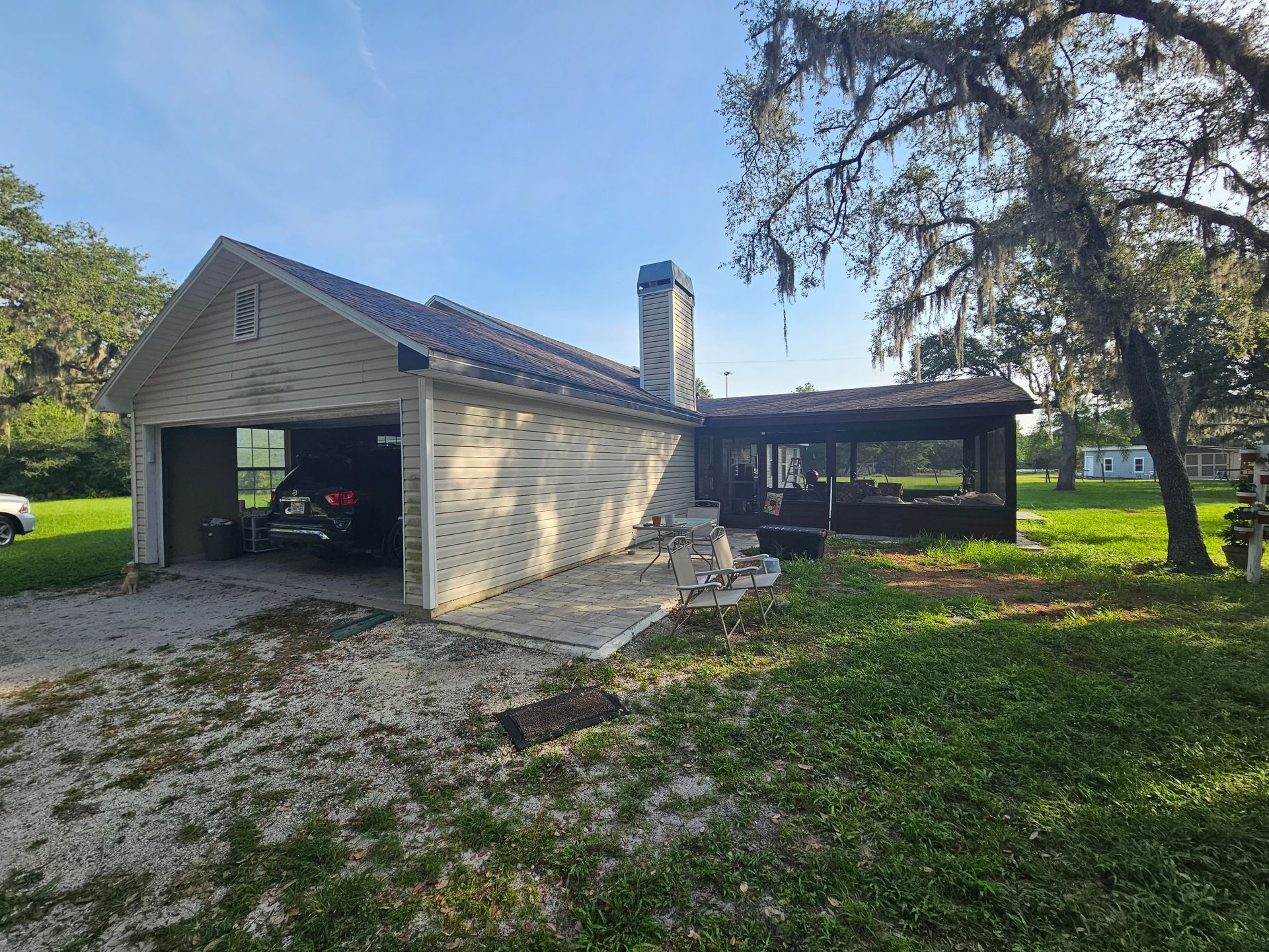 Single-story beige house with open garage, screened porch, and large trees on a grassy yard