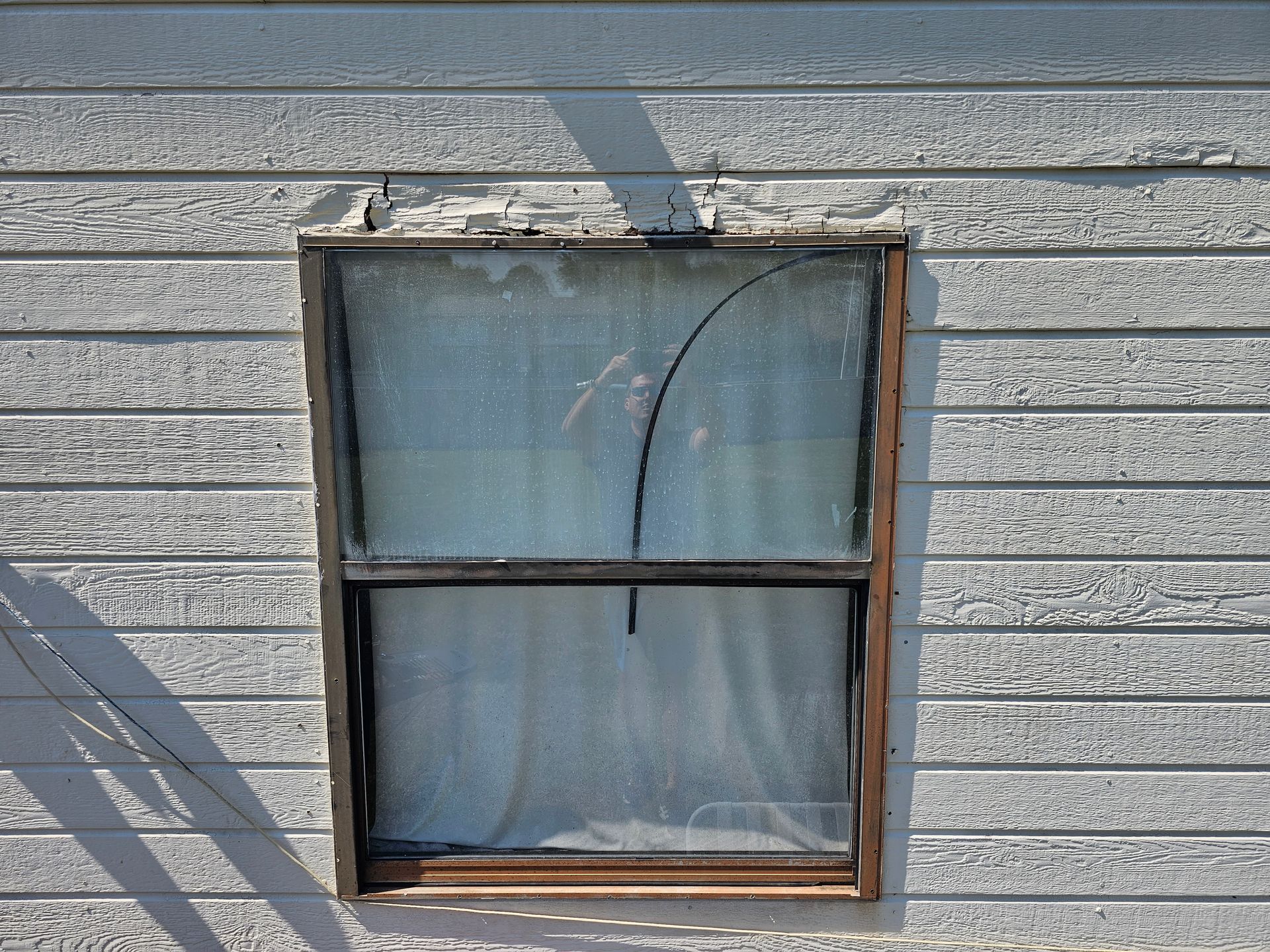 Small weathered window with blue curtains on gray wood siding, sunlight casting shadows.