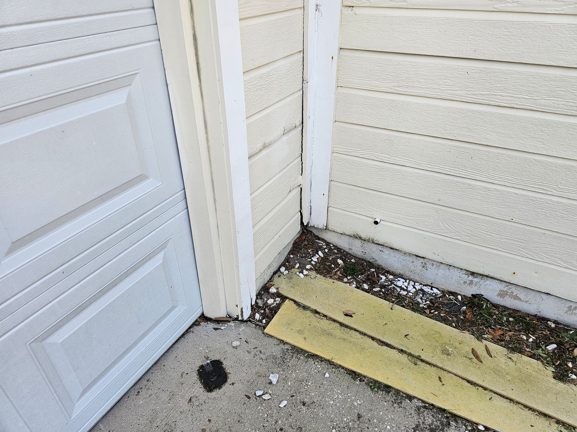 Corner of a garage with white siding, a yellow board on the ground, and debris near the door gap.
