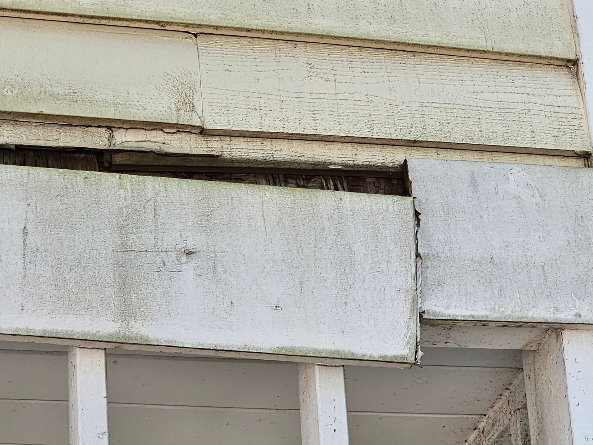 Weathered white house siding with a damaged, misaligned exterior panel and visible gap above a window.