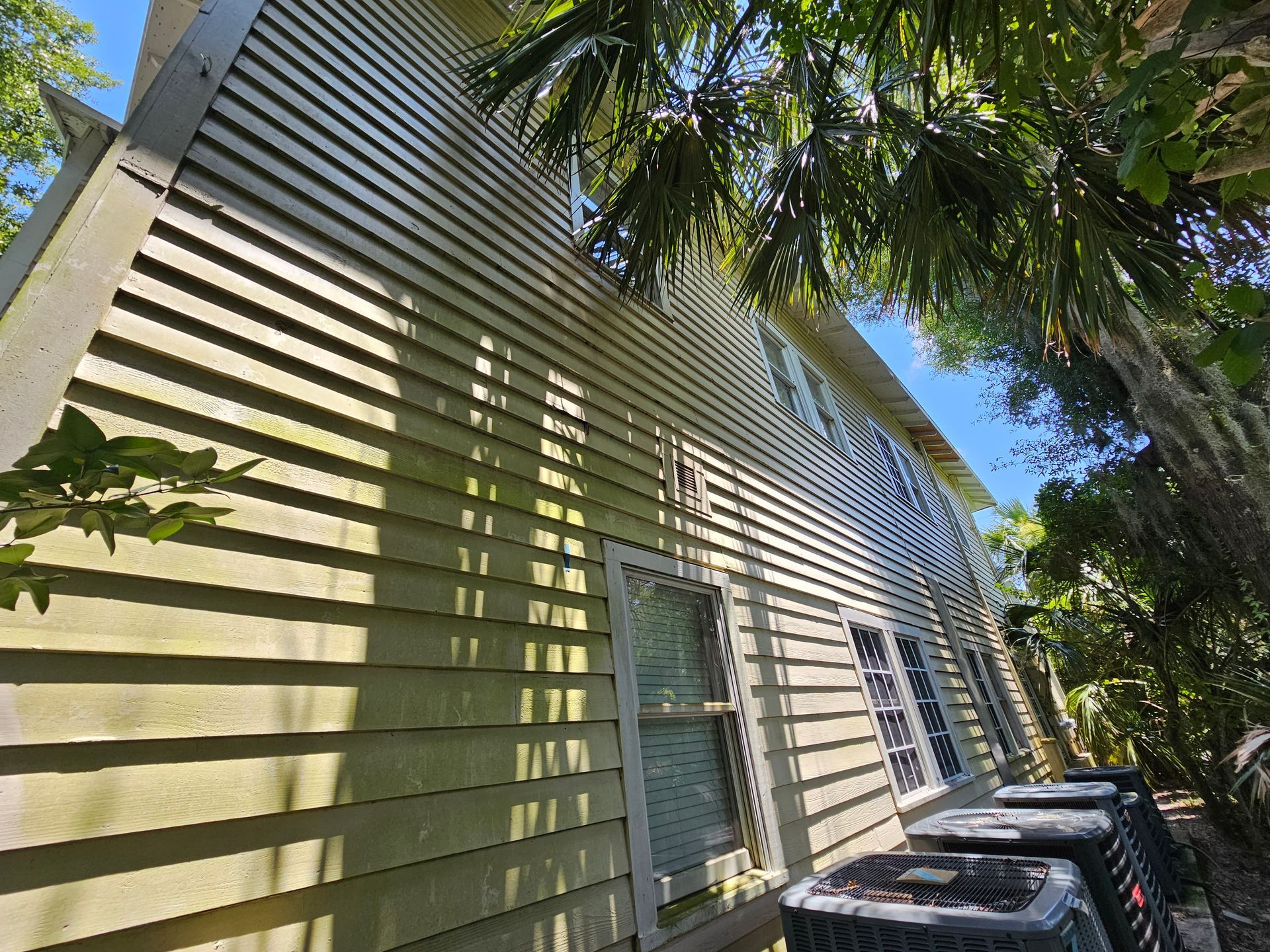 Side of a beige house with windows, tree shadows, and a row of trash bins along the wall.