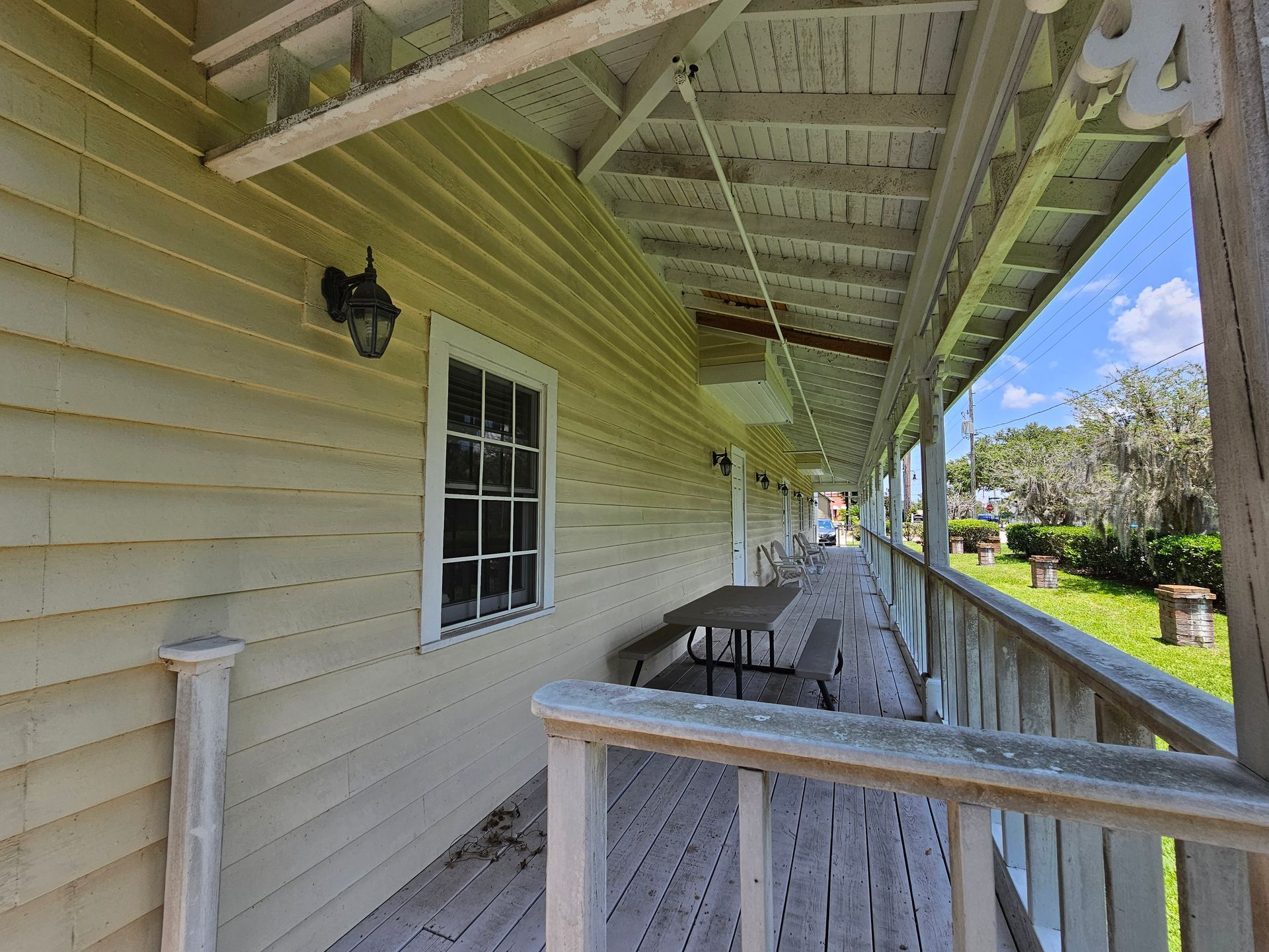 Covered porch with beige siding, wooden railing, bench, and bright blue sky beyond the yard