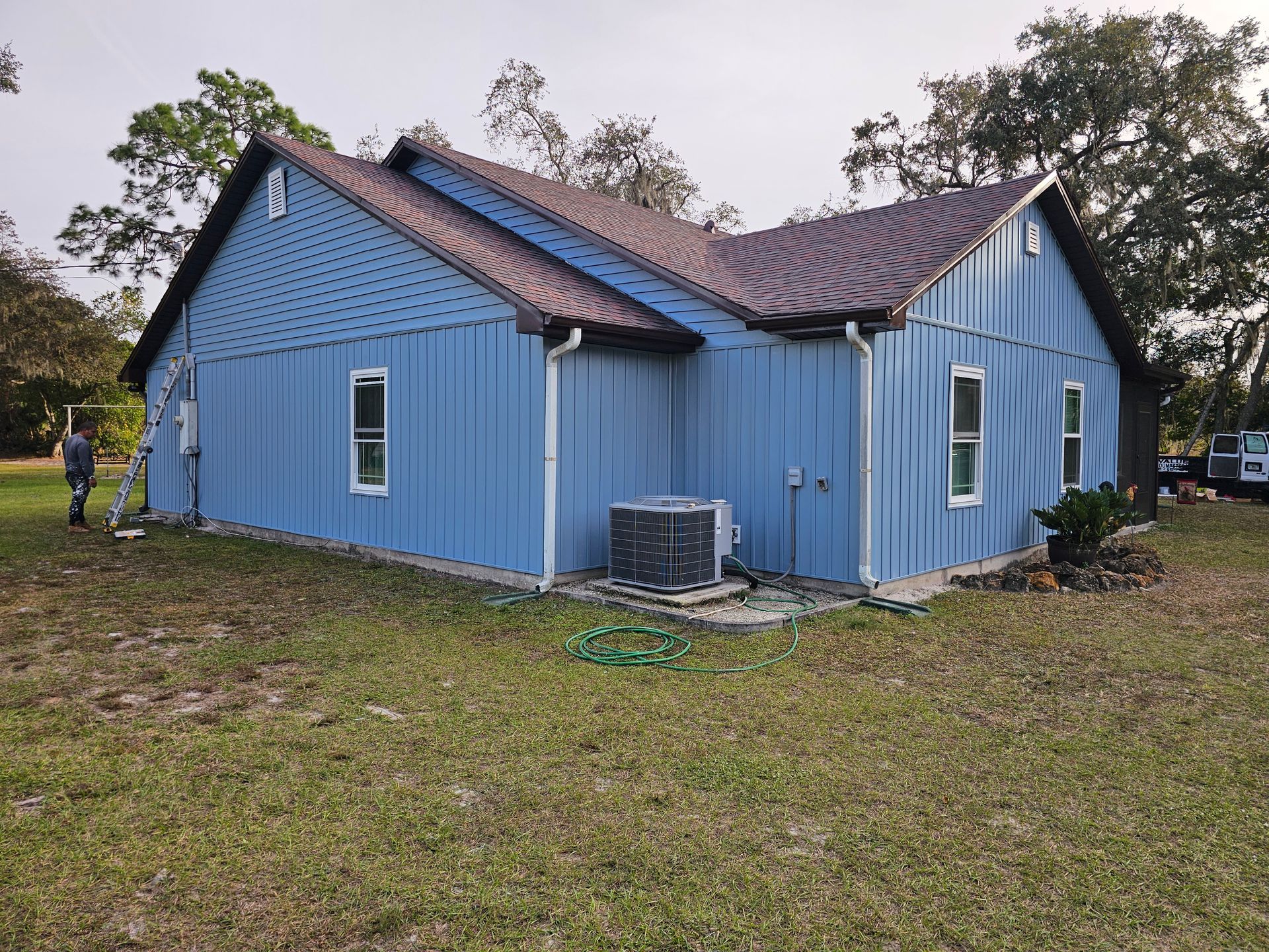 Blue house with damaged roof and siding in a grassy yard, with trees in the background