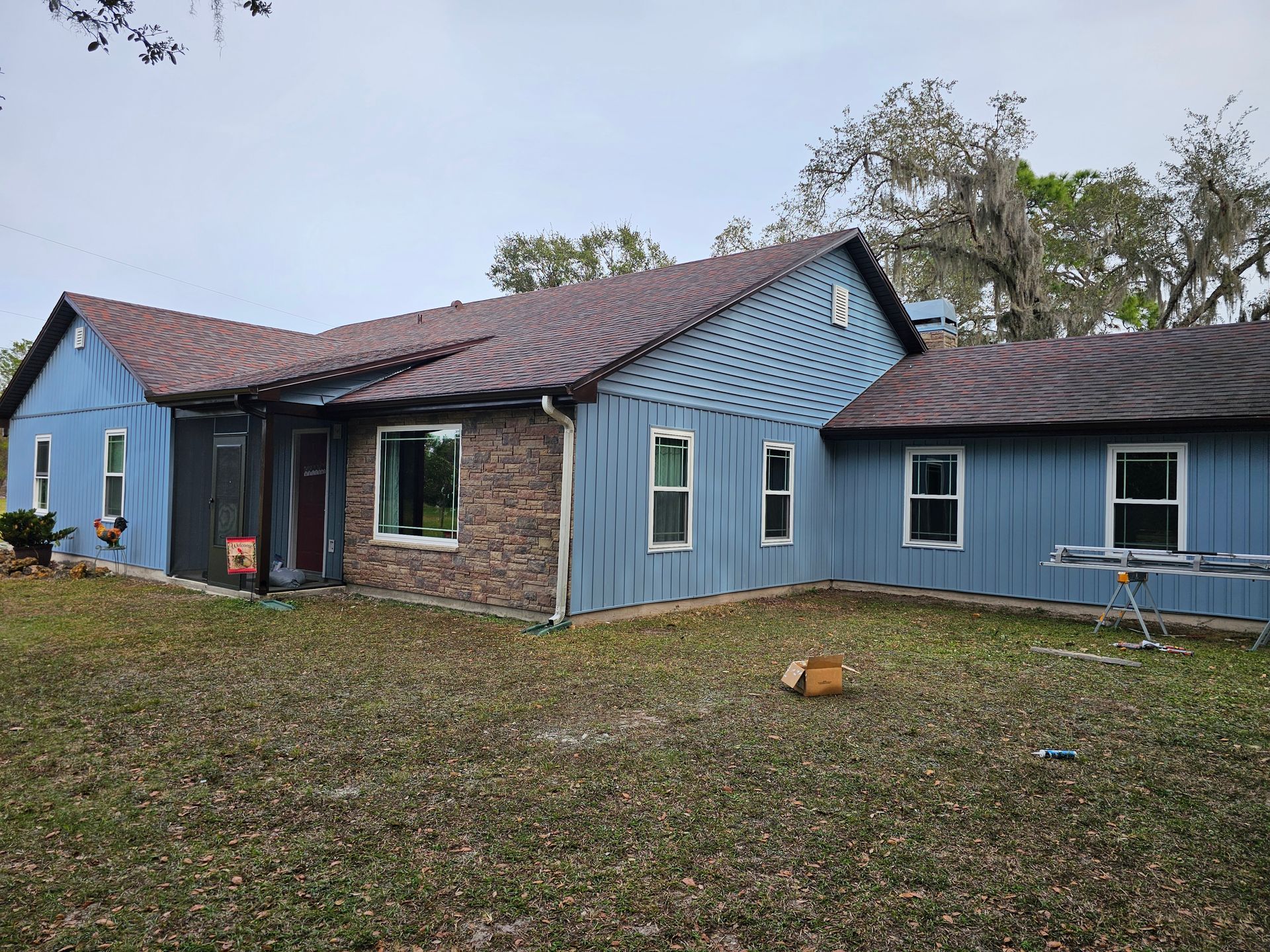 Blue house with a brick center section, dark roof, and grassy yard under a cloudy sky