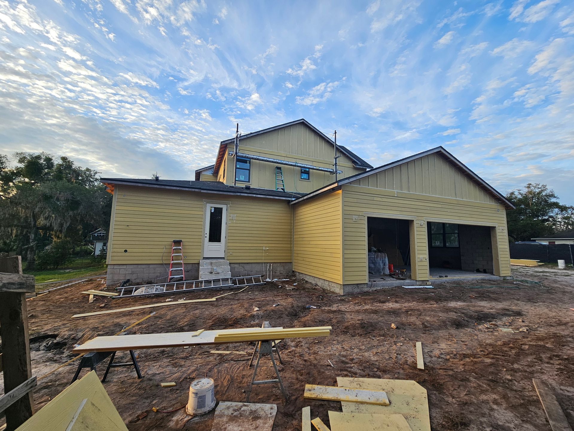 Yellow house under construction with open garage and rough dirt yard beneath a cloudy sky