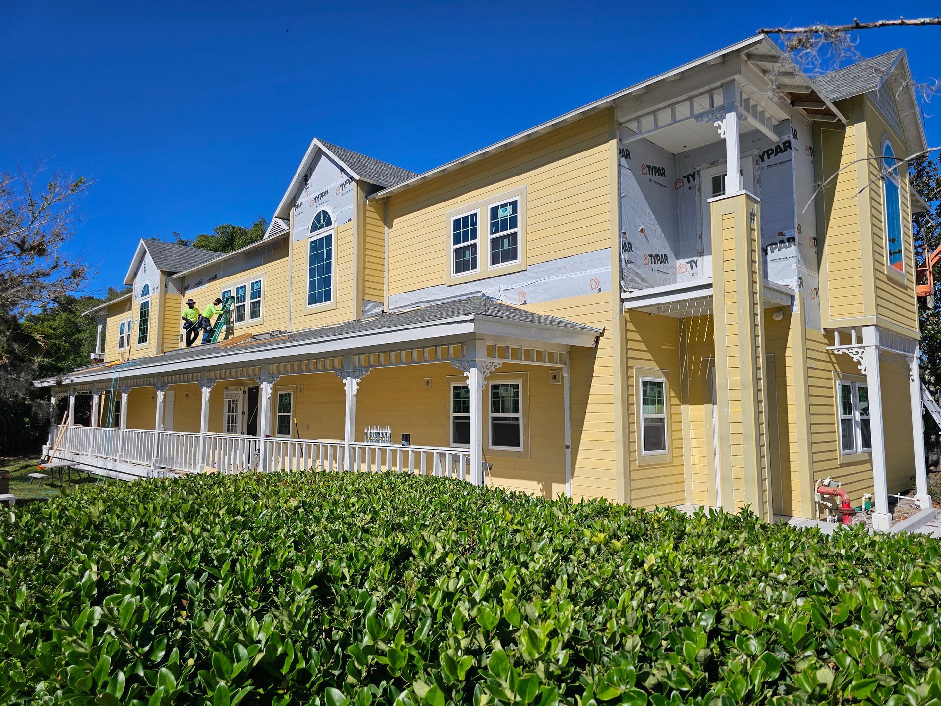 Yellow two-story building with white trim and covered porch beside green hedges under a blue sky