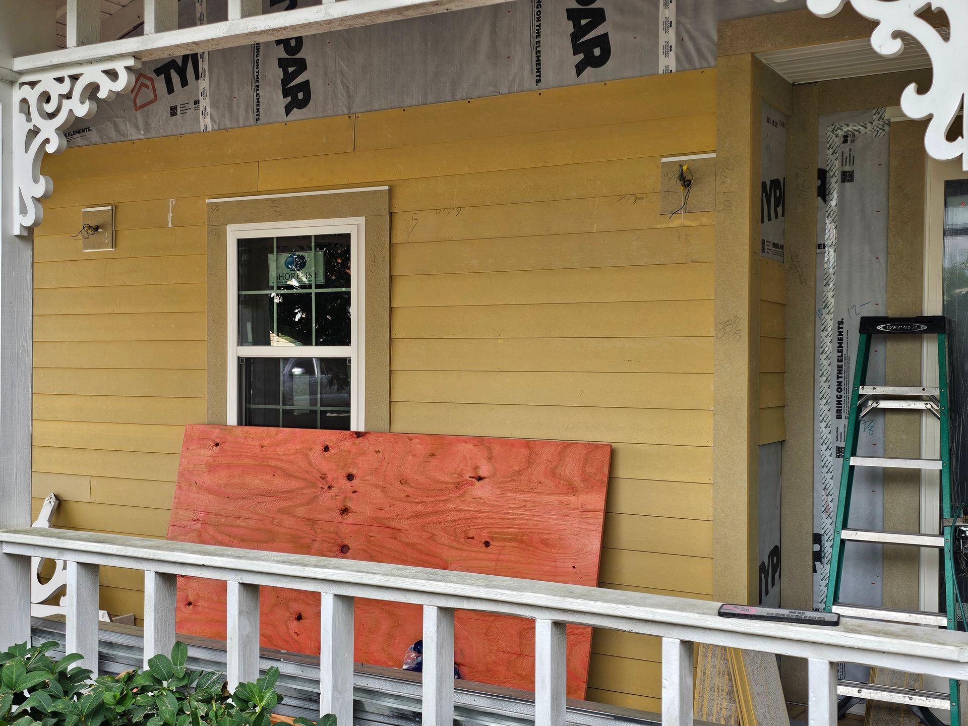 Yellow house porch under renovation with plywood-covered railing, window, and a ladder on the right
