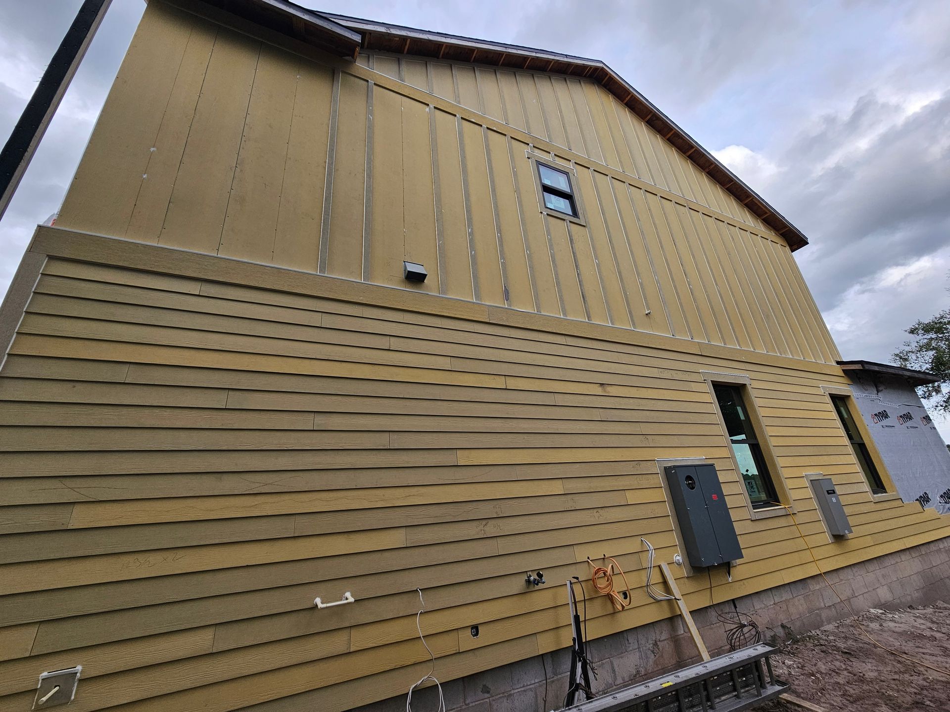 Side view of a tan building with horizontal siding, small windows, utility boxes, and gravel ground under cloudy sky