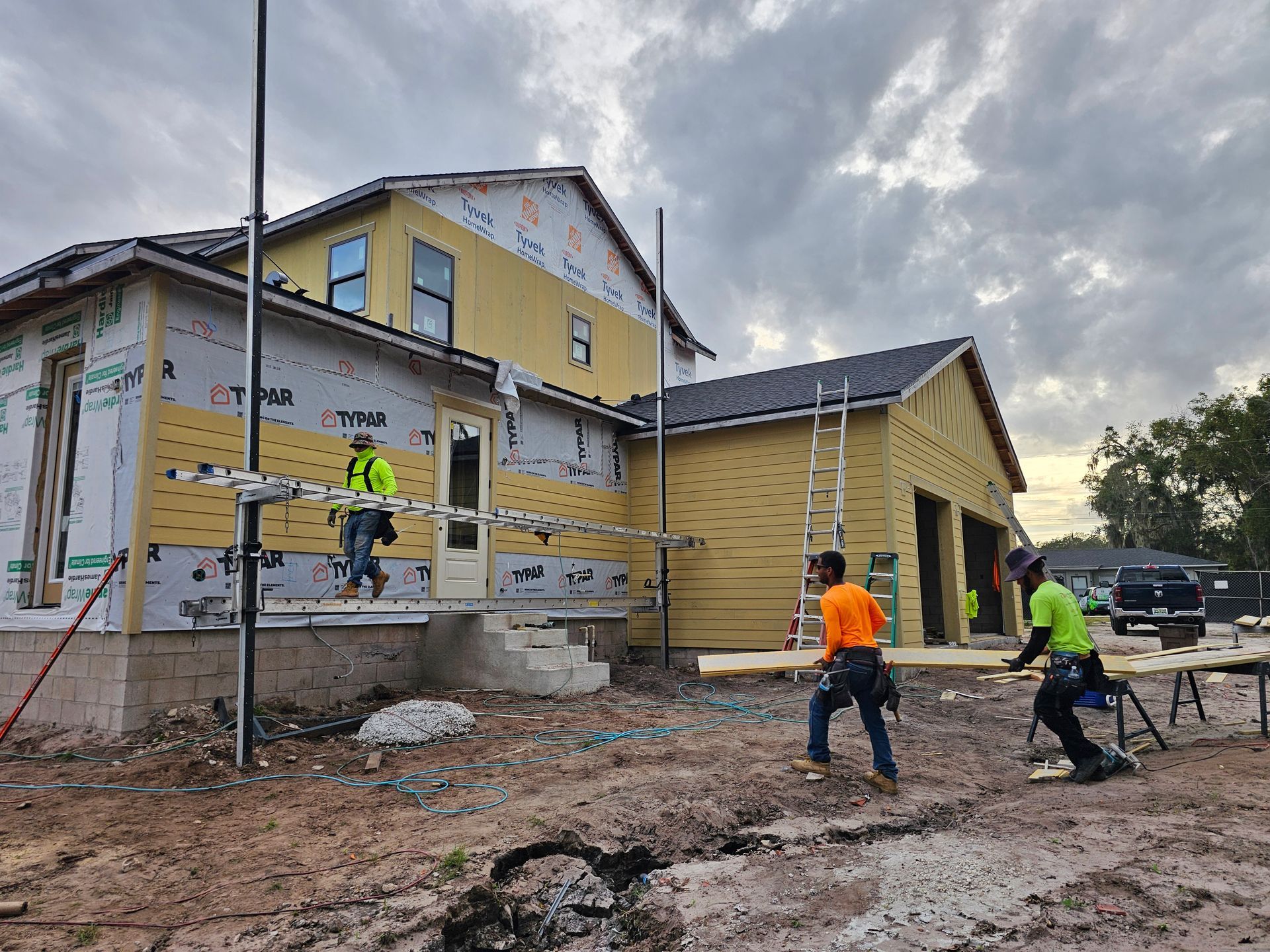 Construction workers in bright vests install house siding on a muddy residential build site under cloudy skies
