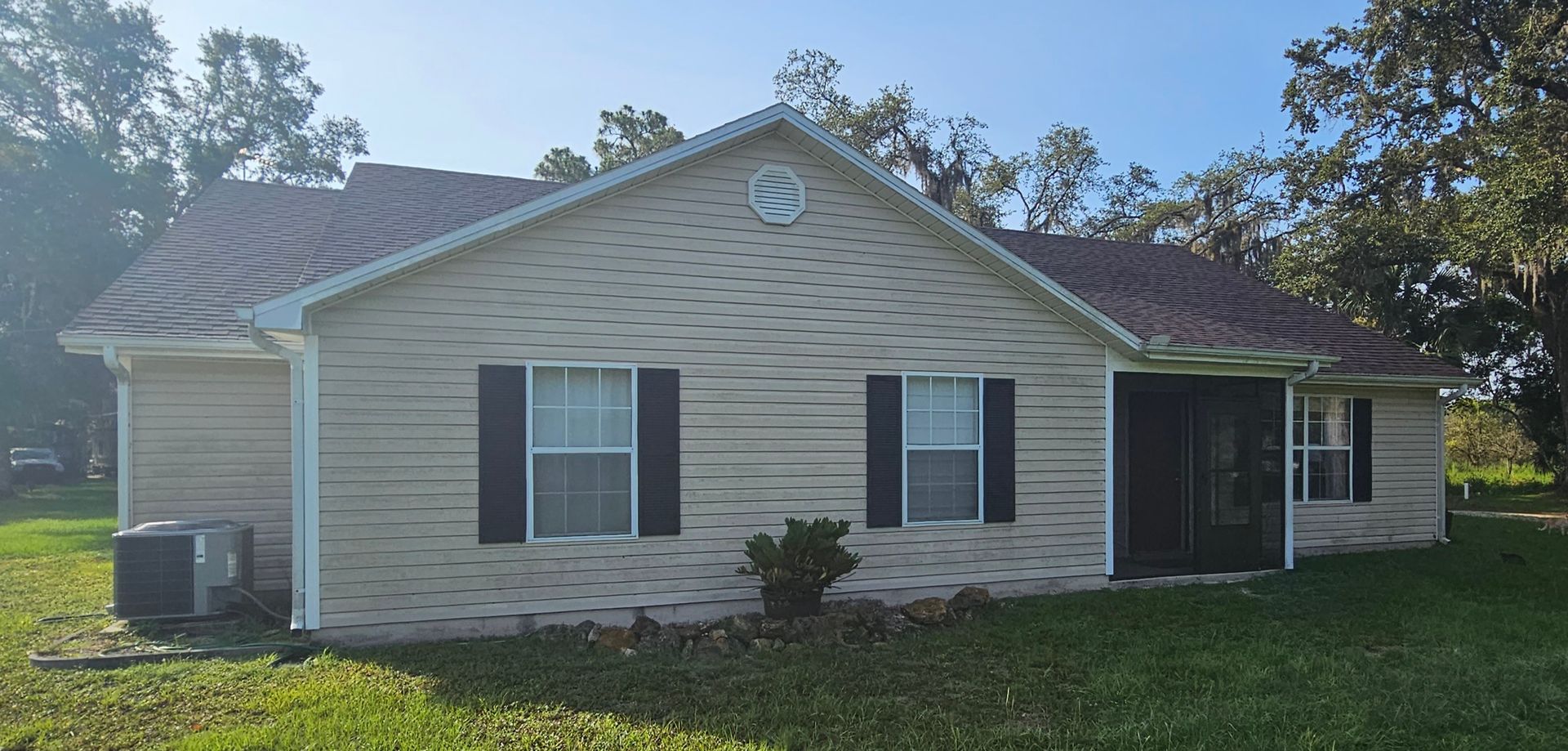 Single-story beige house with black shutters and a small covered porch, surrounded by grass and trees