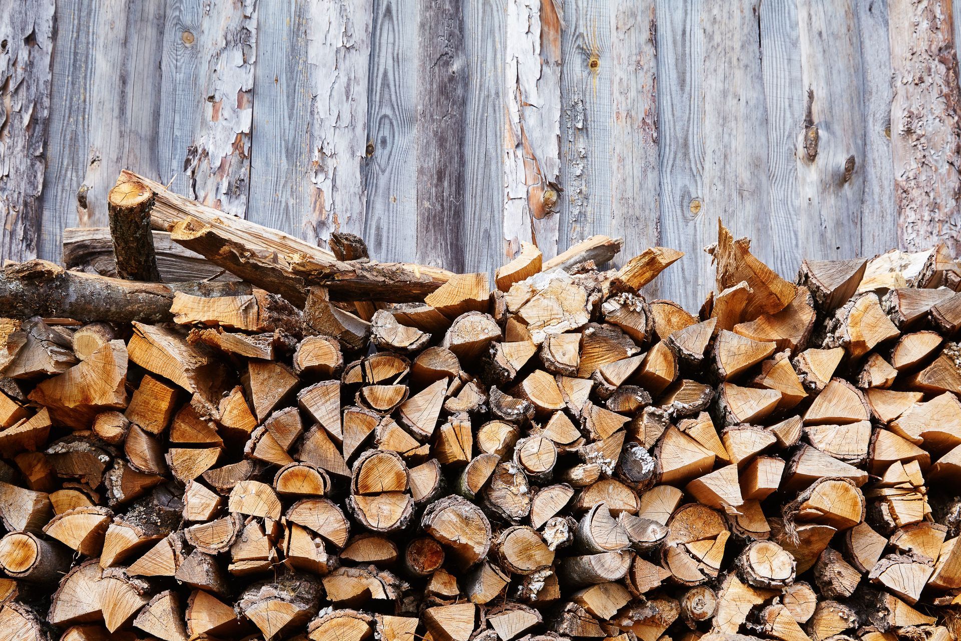 Pile of split firewood in front of weathered, gray wooden wall.