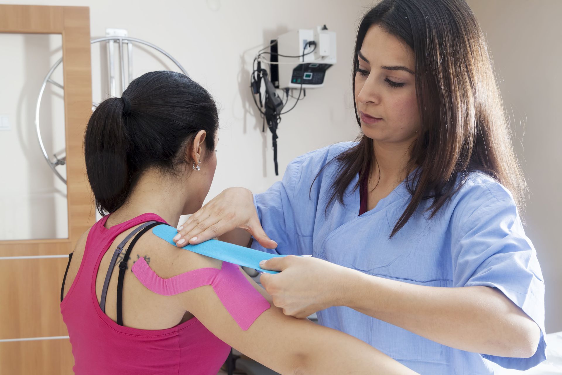 Healthcare worker applying blue kinesiology tape to a patient&rsquo;s pink shoulder in a clinic room