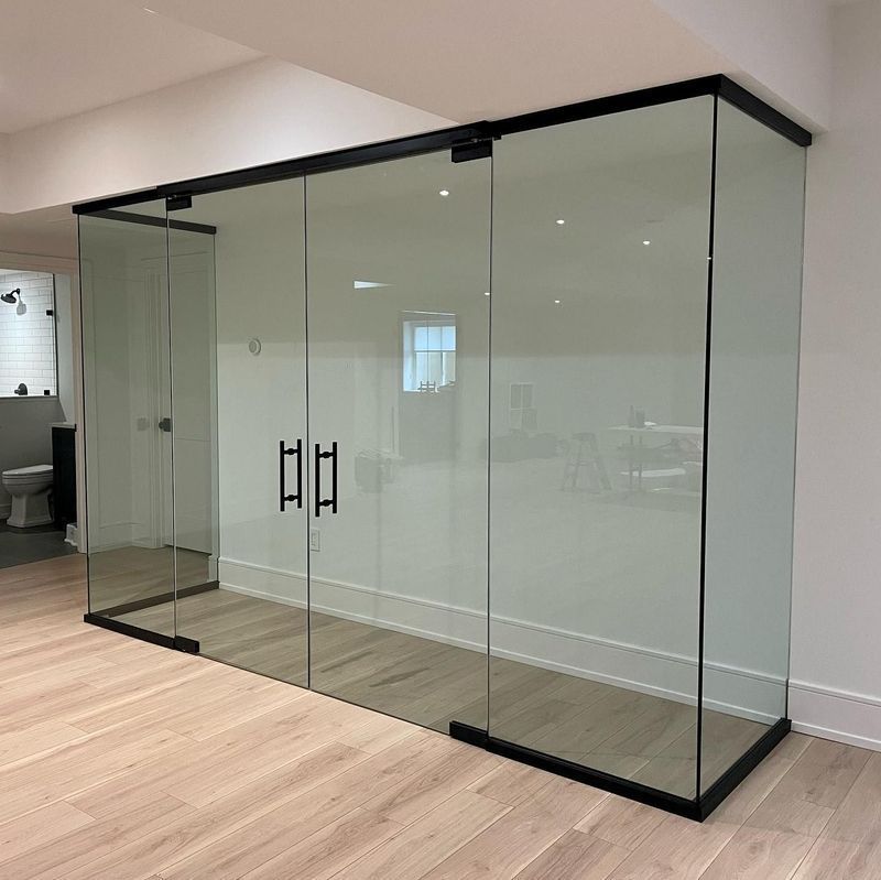 Glass-walled room with black frame, wooden floor, and black door handles. Interior view of a bathroom visible through the glass.