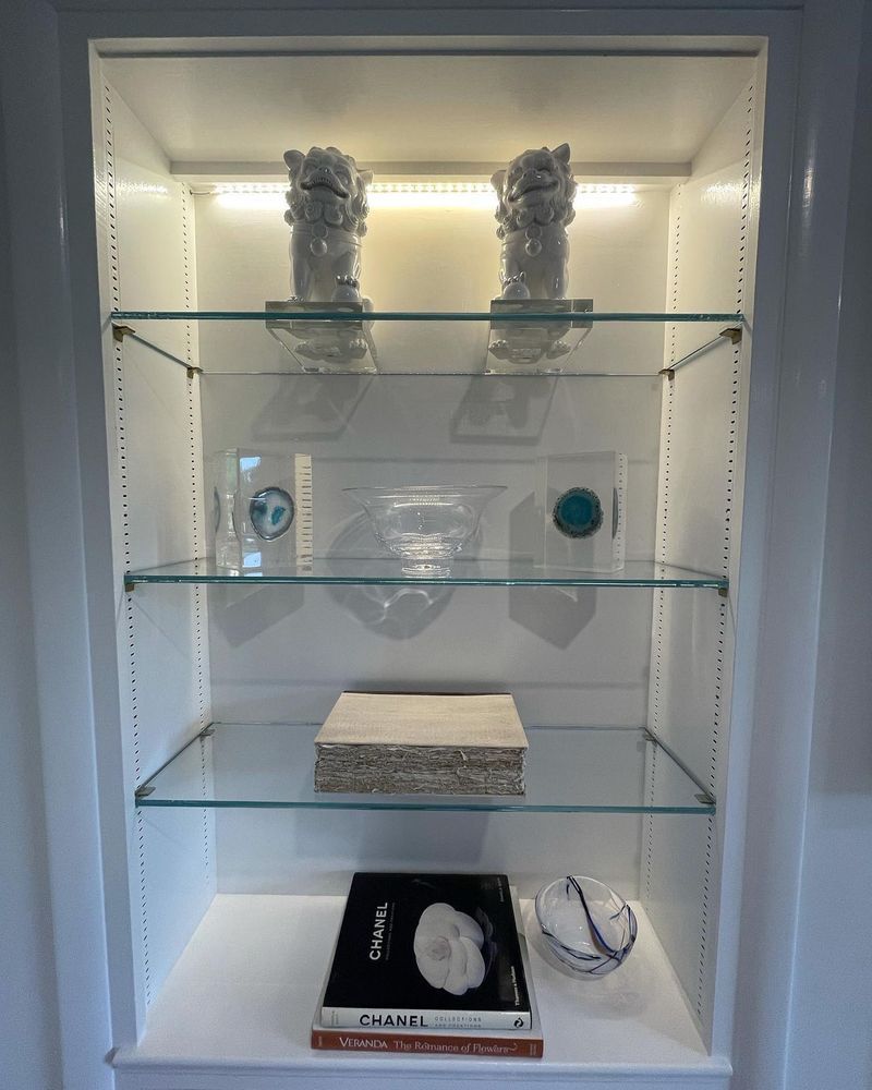 White display cabinet with glass shelves, showcasing decorative objects, lit with overhead light.