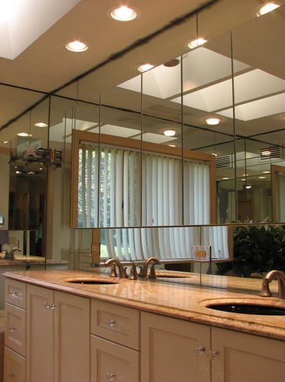 Bathroom with mirrored walls, double sinks, light-colored cabinets, and a window with blinds.