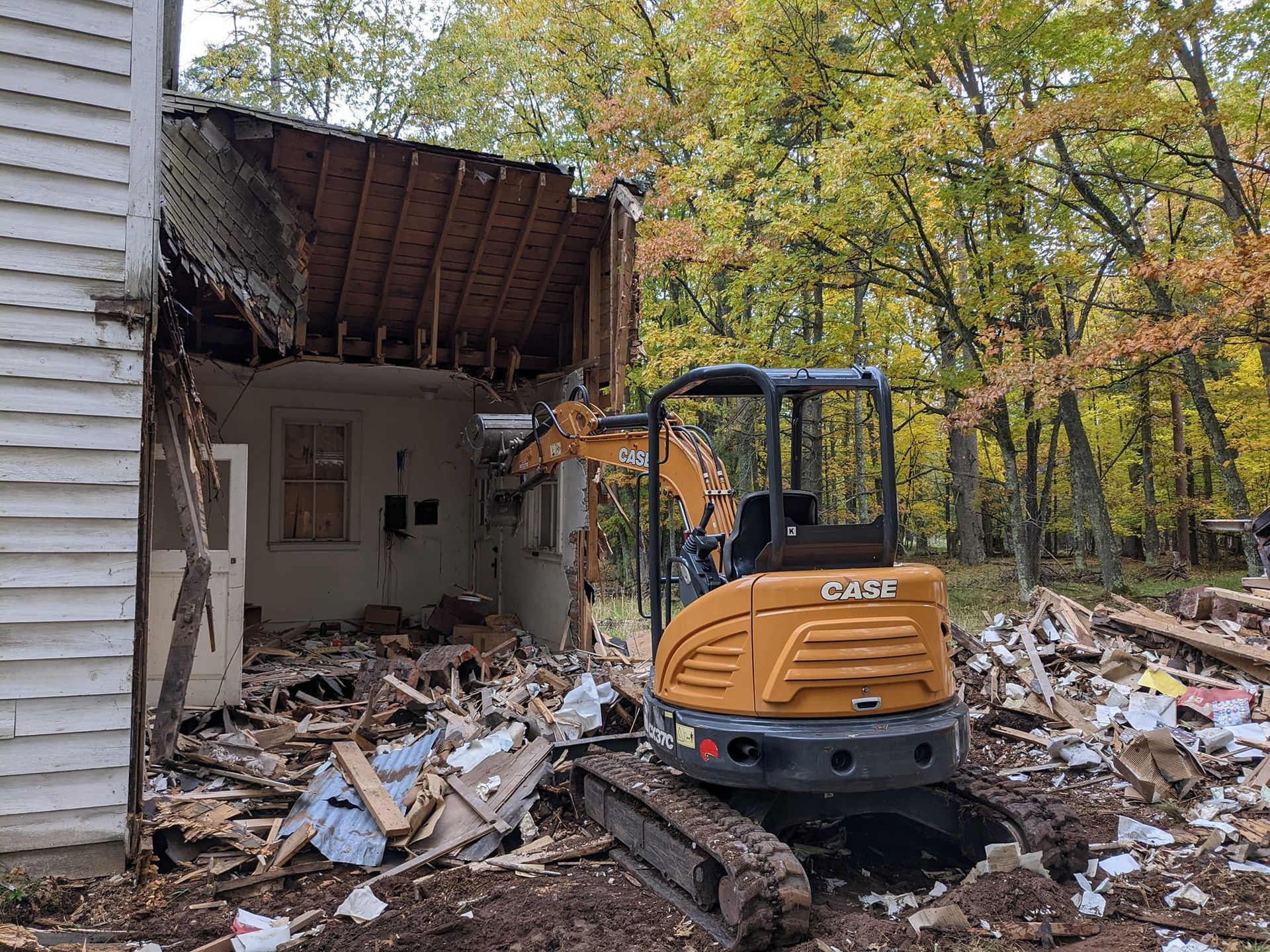 A small excavator is demolishing a house in the woods.