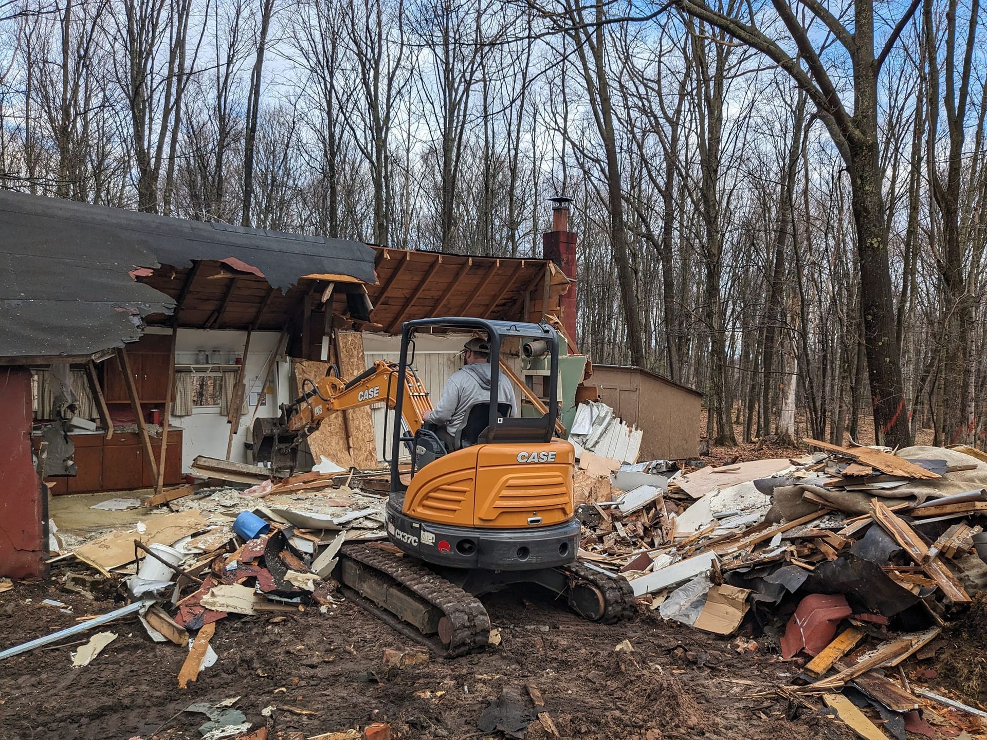 A small excavator is demolishing a house in the woods.