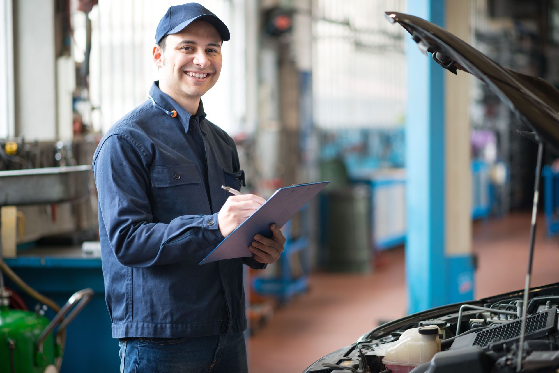 Mechanic smiling and writing on a clipboard beside an open car hood in a workshop
