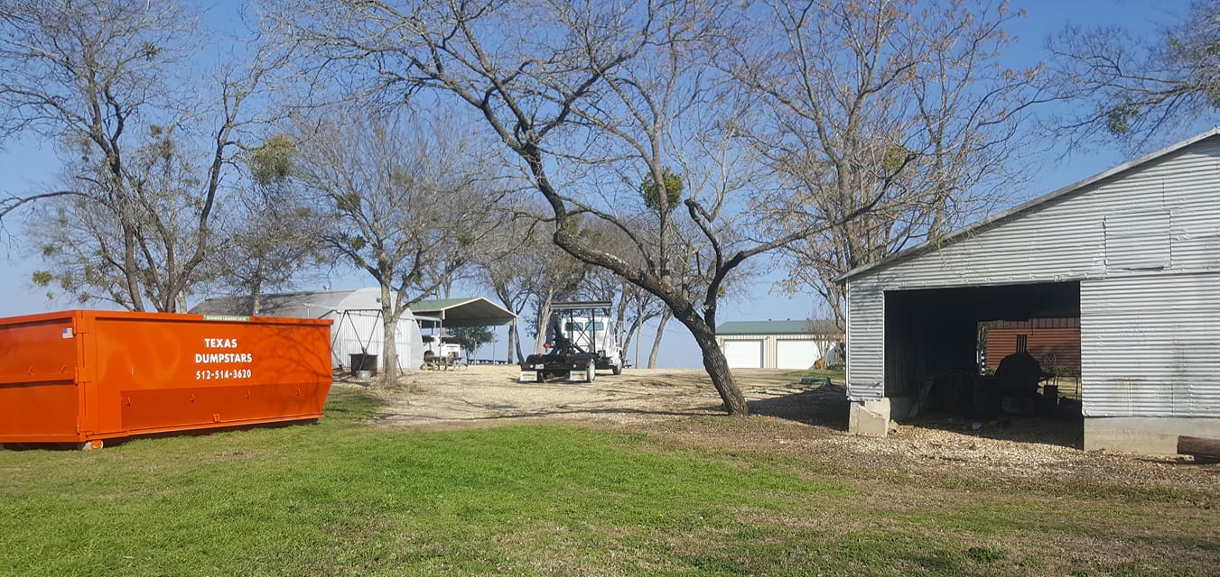 A large orange dumpster is sitting in a grassy field next to a garage.