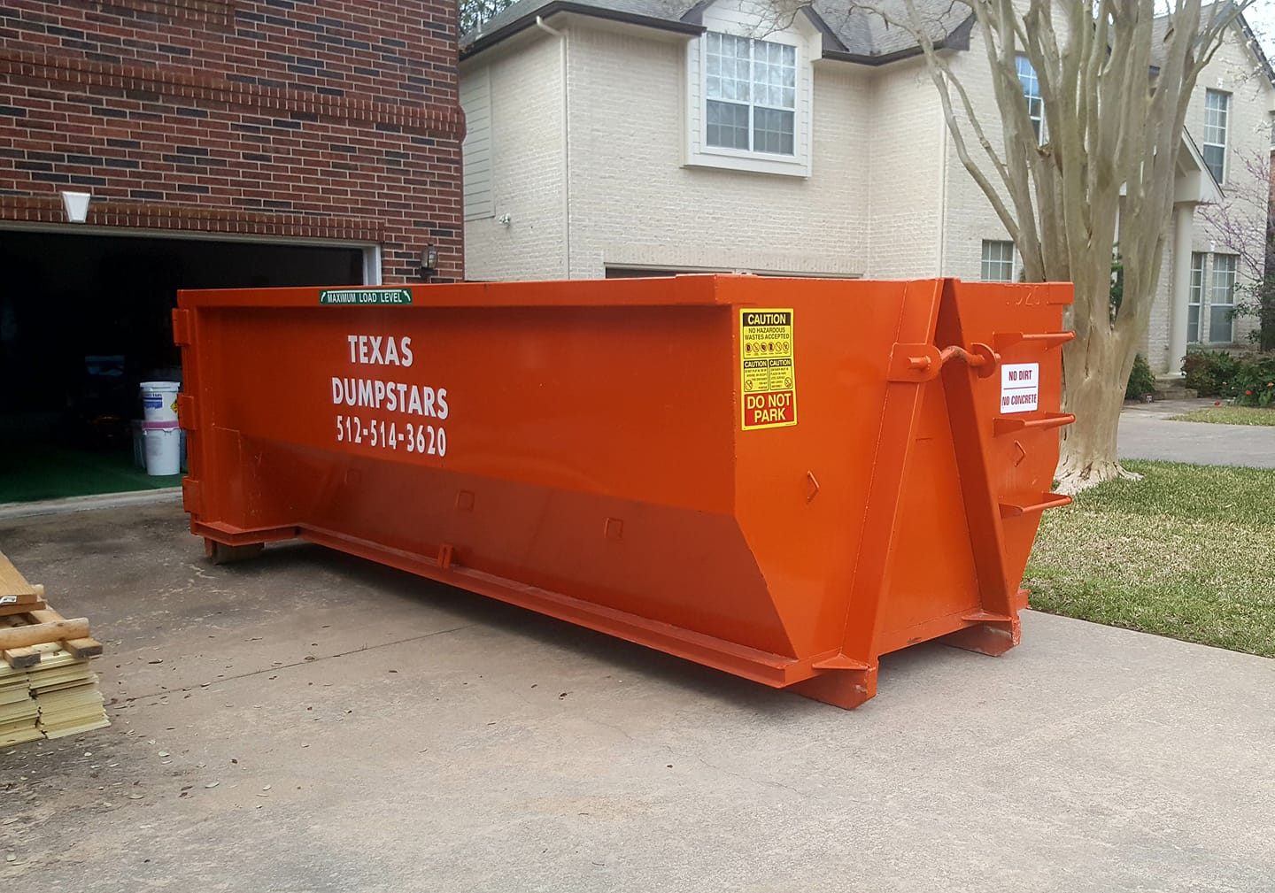 A large orange dumpster is parked in front of a brick house.