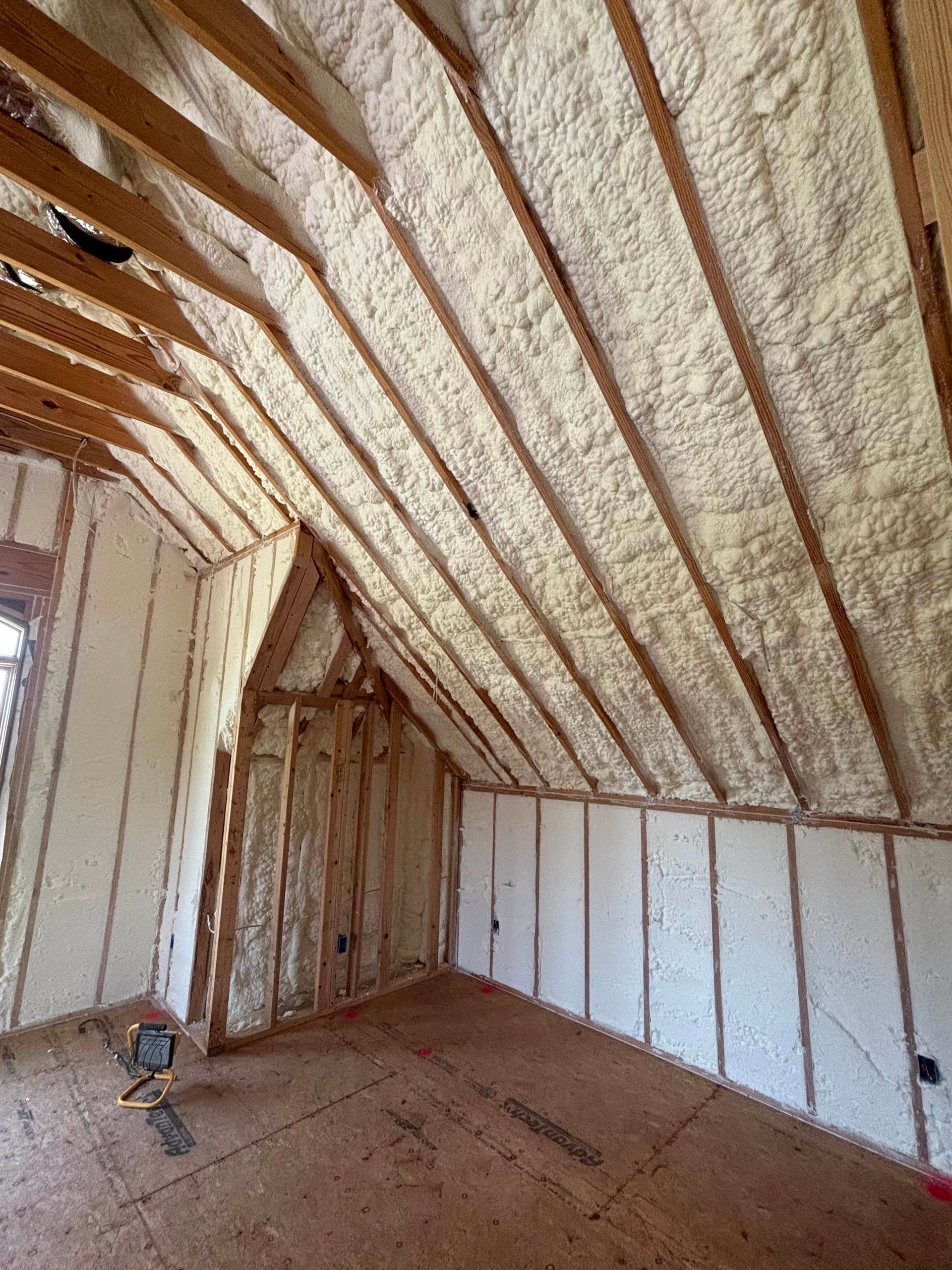 Attic room interior under construction with spray foam insulation applied between wooden wall and ceiling rafters.