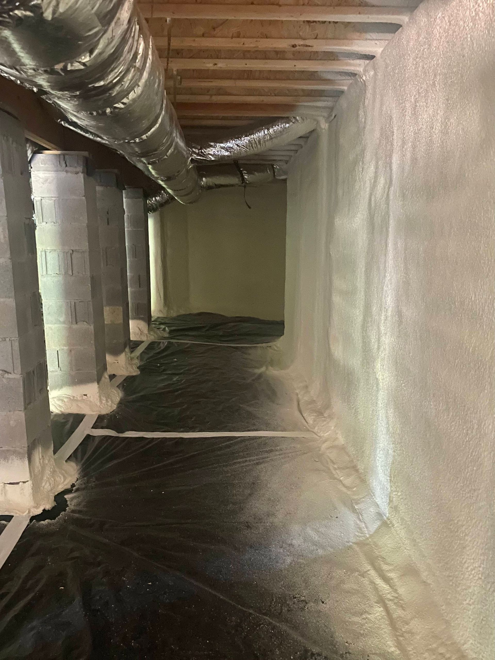 A crawl space with foam-insulated concrete walls, a dark vapor barrier on the ground, and overhead ductwork.