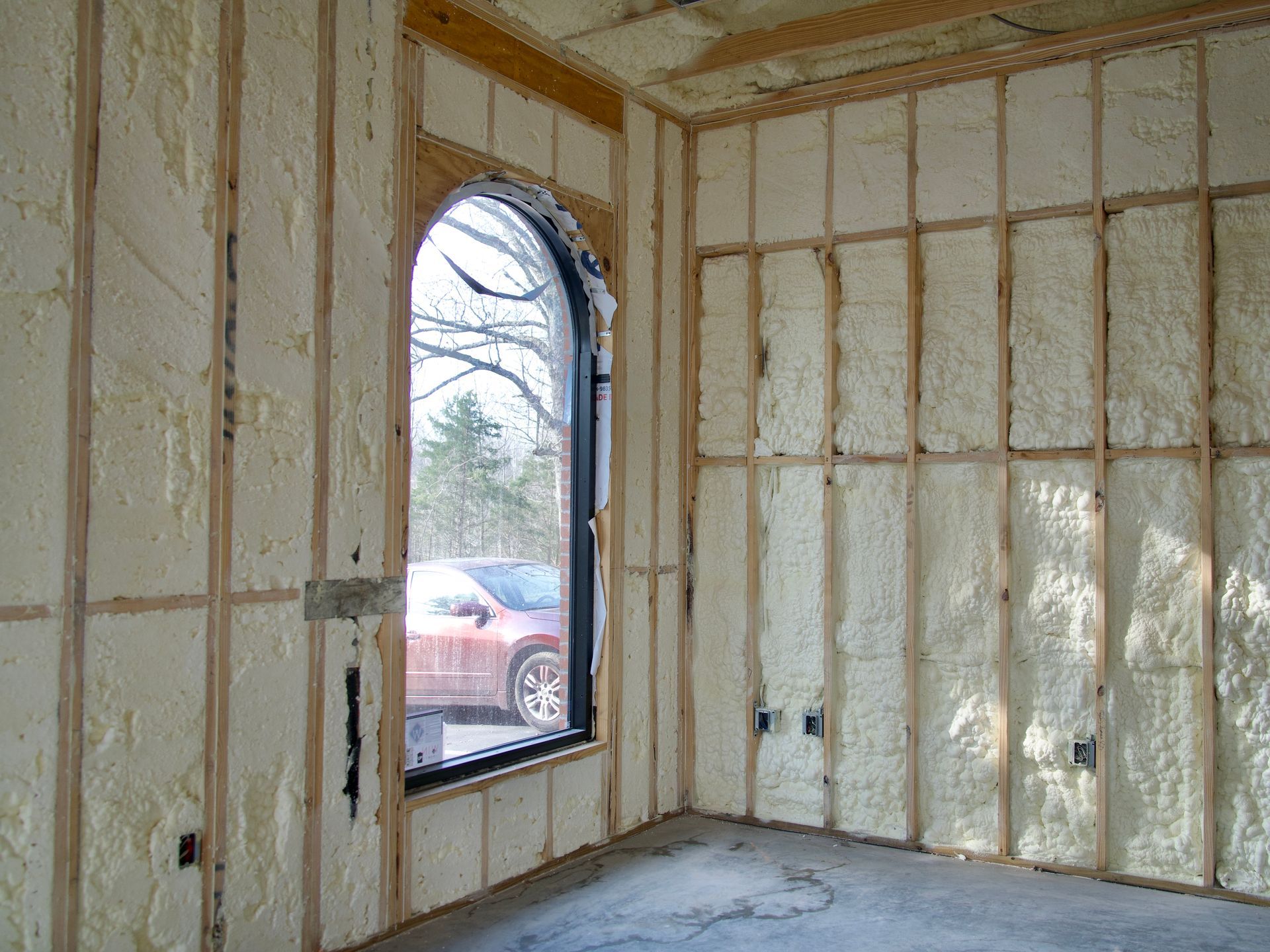 Interior view of unfinished wood wall studs filled with light yellow spray foam insulation and an arched window.